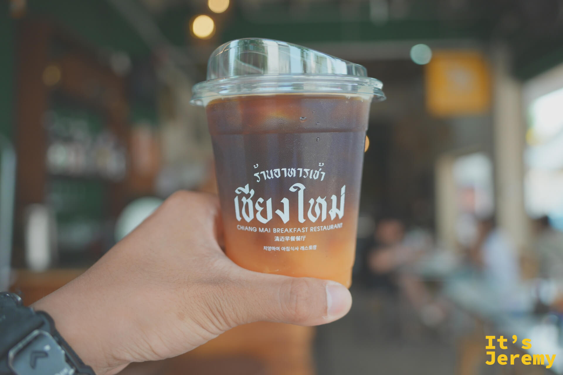 手持印有清邁早餐餐廳 Logo 的 Orange Americano 透明杯,背景店內空間