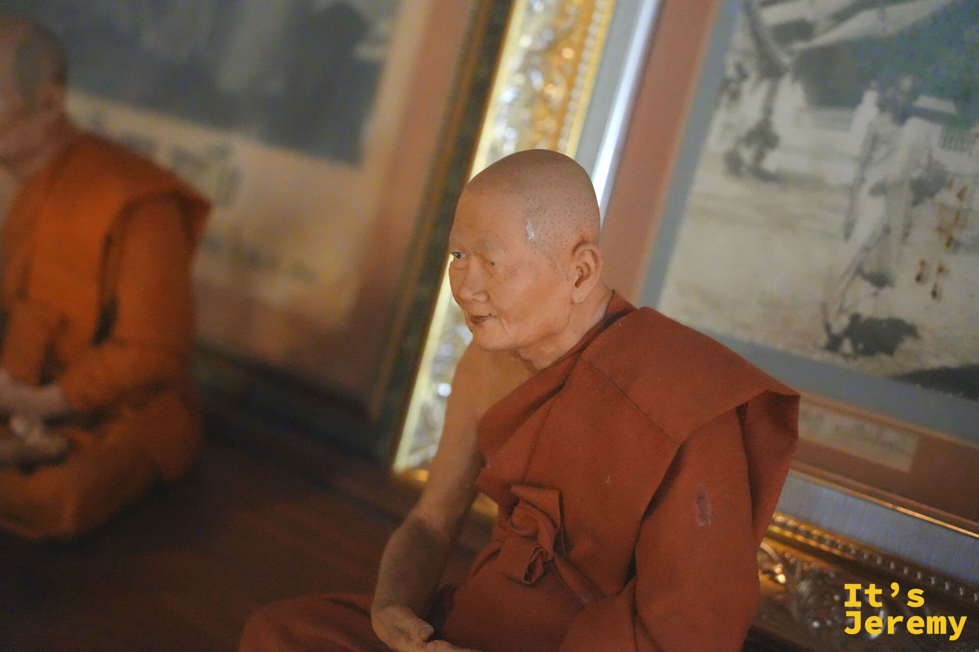 高僧蠟像,面部細節高度逼真,穿著橘色僧衣,室內光線昏暗,氛圍肅穆