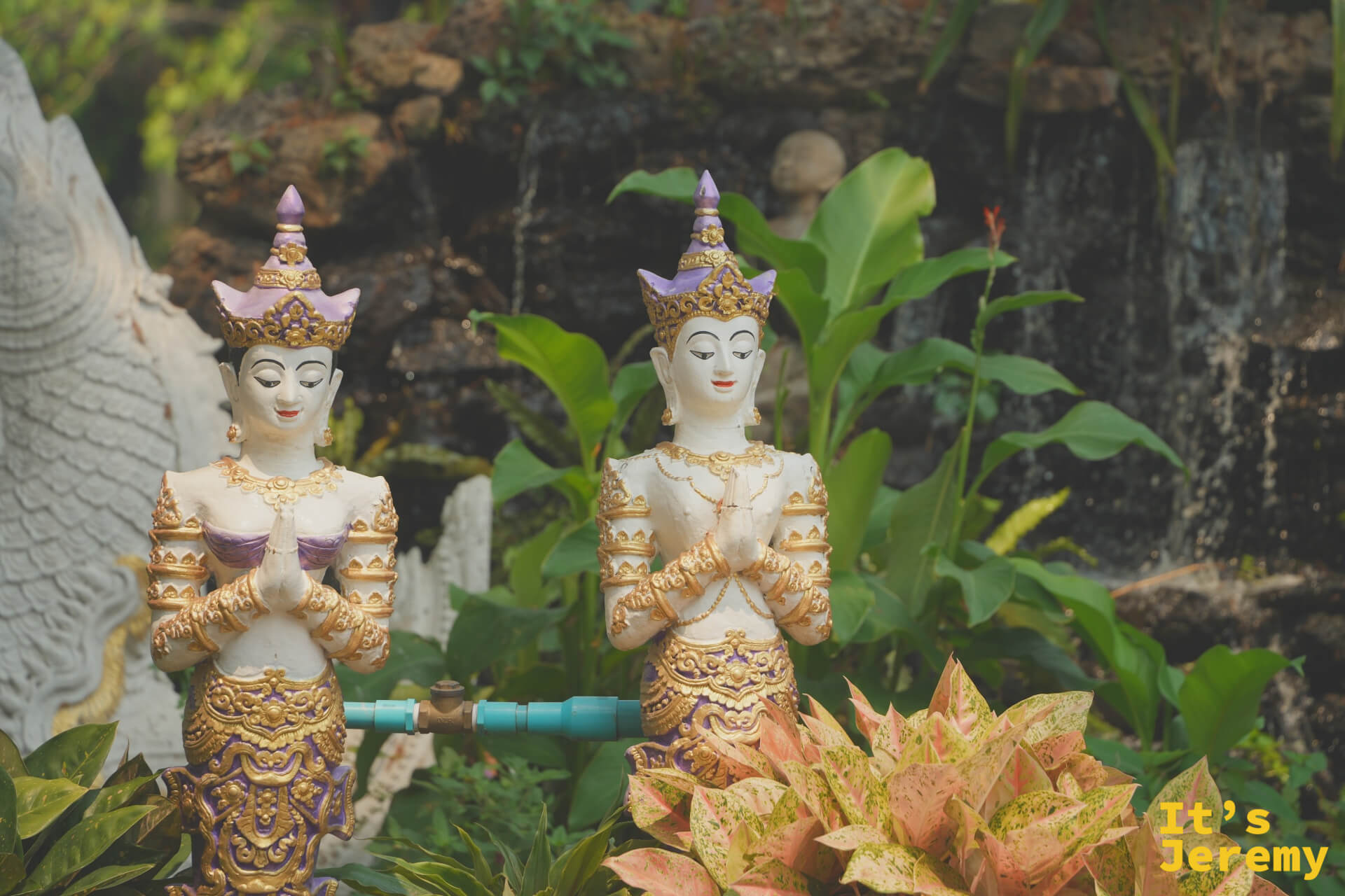 兩尊紫金配色的天女合掌雕像立於植栽與岩石間，背景有小瀑布流淌，雕工細緻