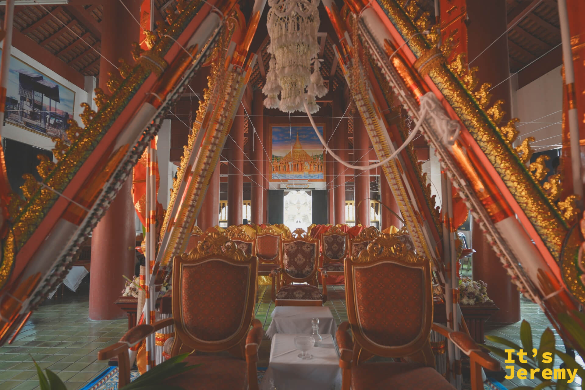 主殿廊柱全景，深棕紅色木柱兩側排開，中央鋪有走道，木椅上覆著紅橙色布料，殿後可見緬甸風格壁畫