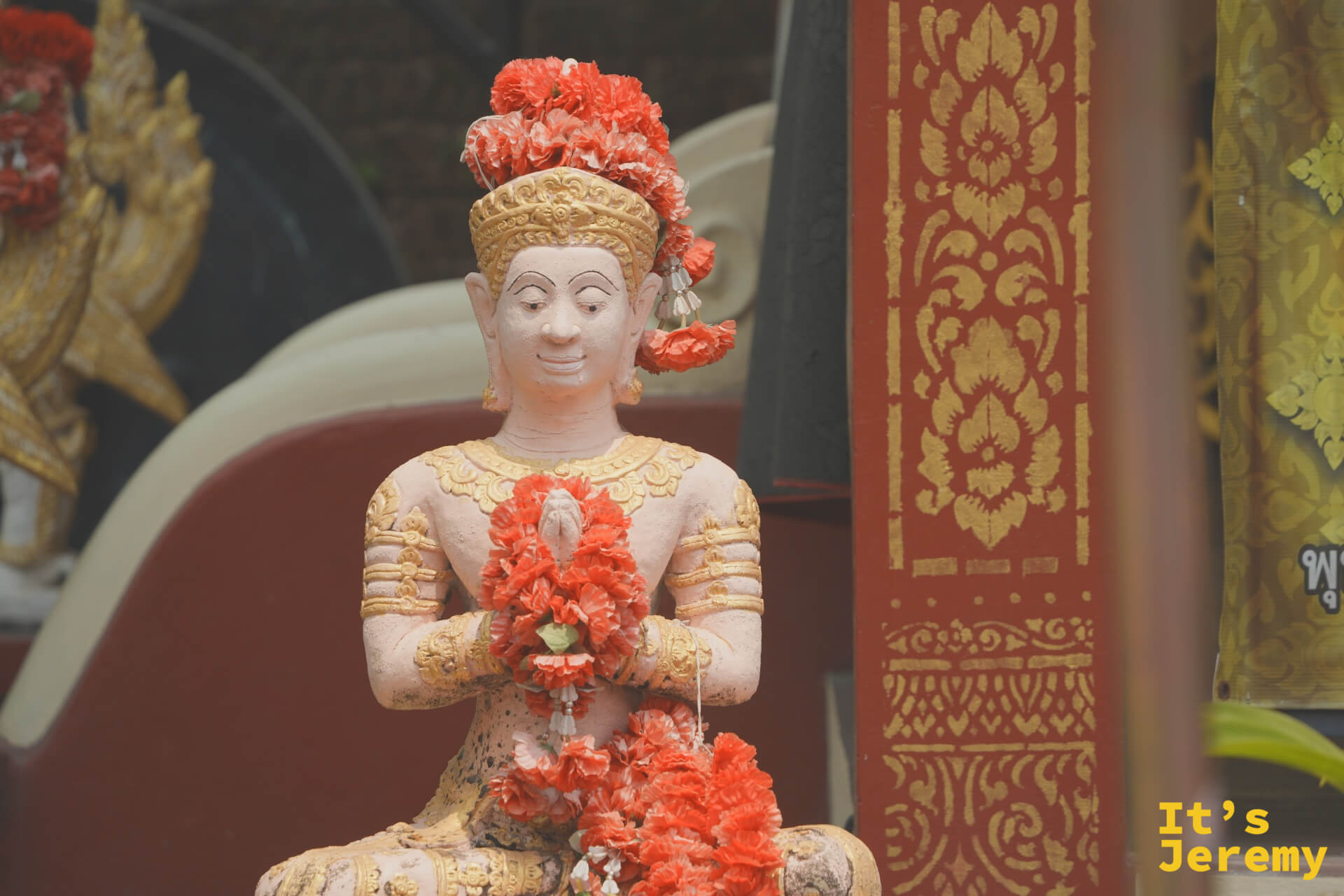 合掌祈願的男性天神像,頭戴紅色花冠,雙手捧著花環,神情專注,背景是金紅色的廟堂裝飾
