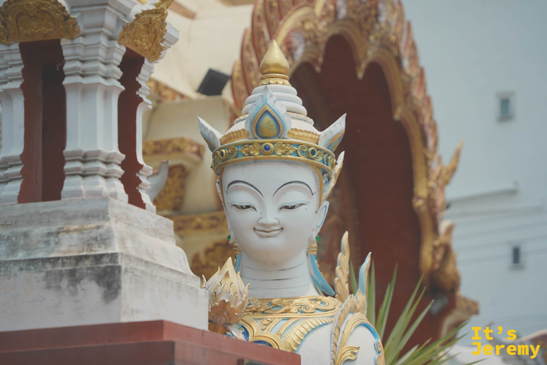 白色金冠的提婆達近景,眼神平靜,藍色瞳仁,後方可見寺廟建築的紅色拱門