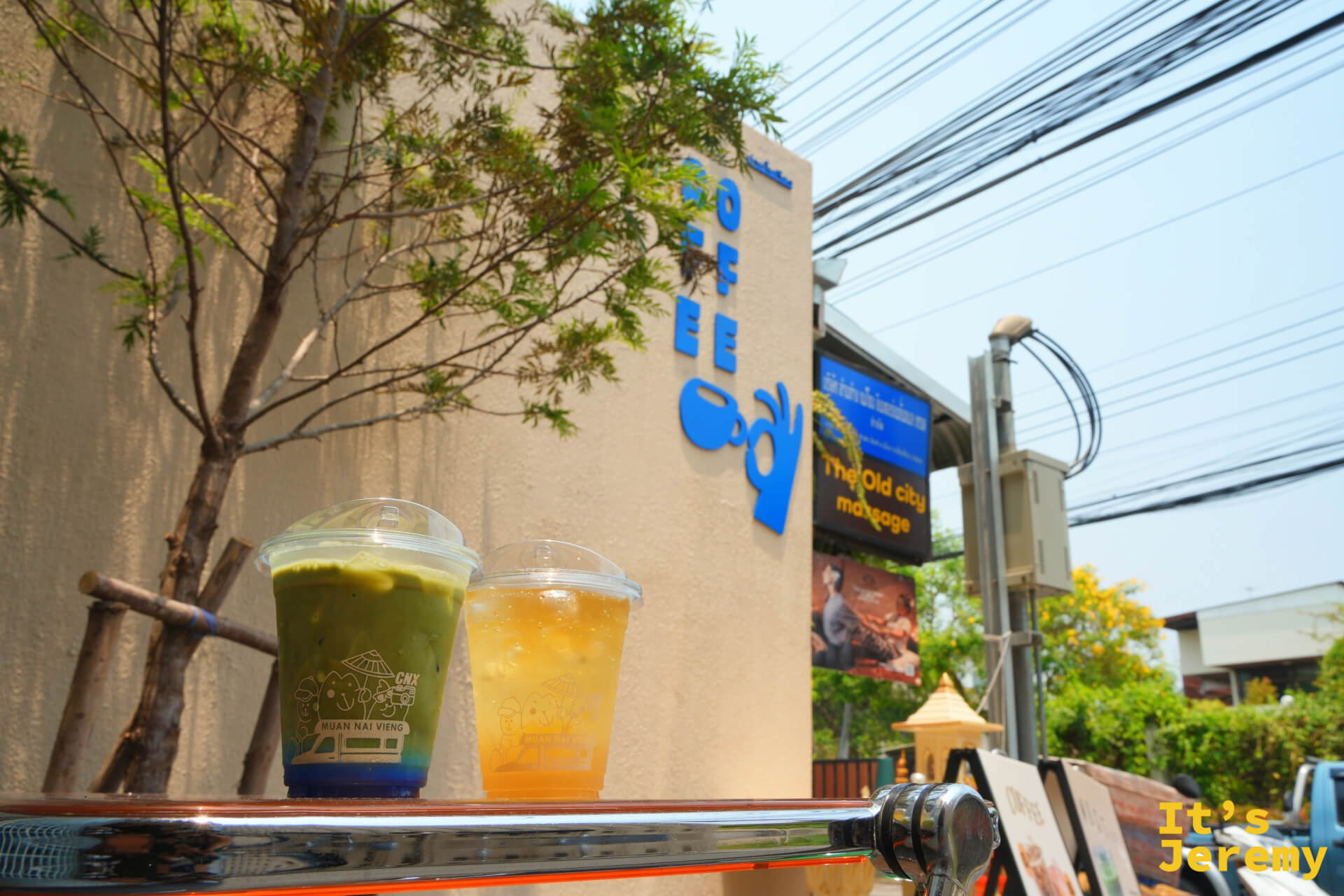 兩杯 Muan Nai Vieng 飲料擺在戶外，背景是藍色 COFFEE 招牌