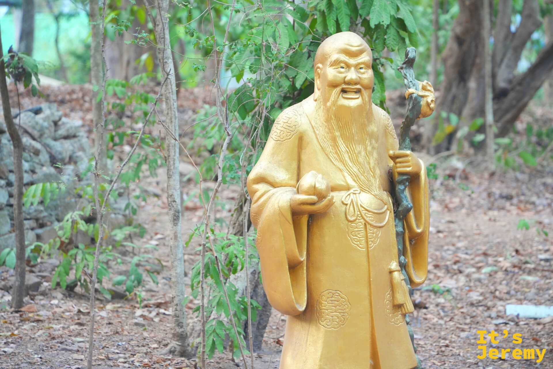 中式老者雕像,持杖微笑,背靠綠樹