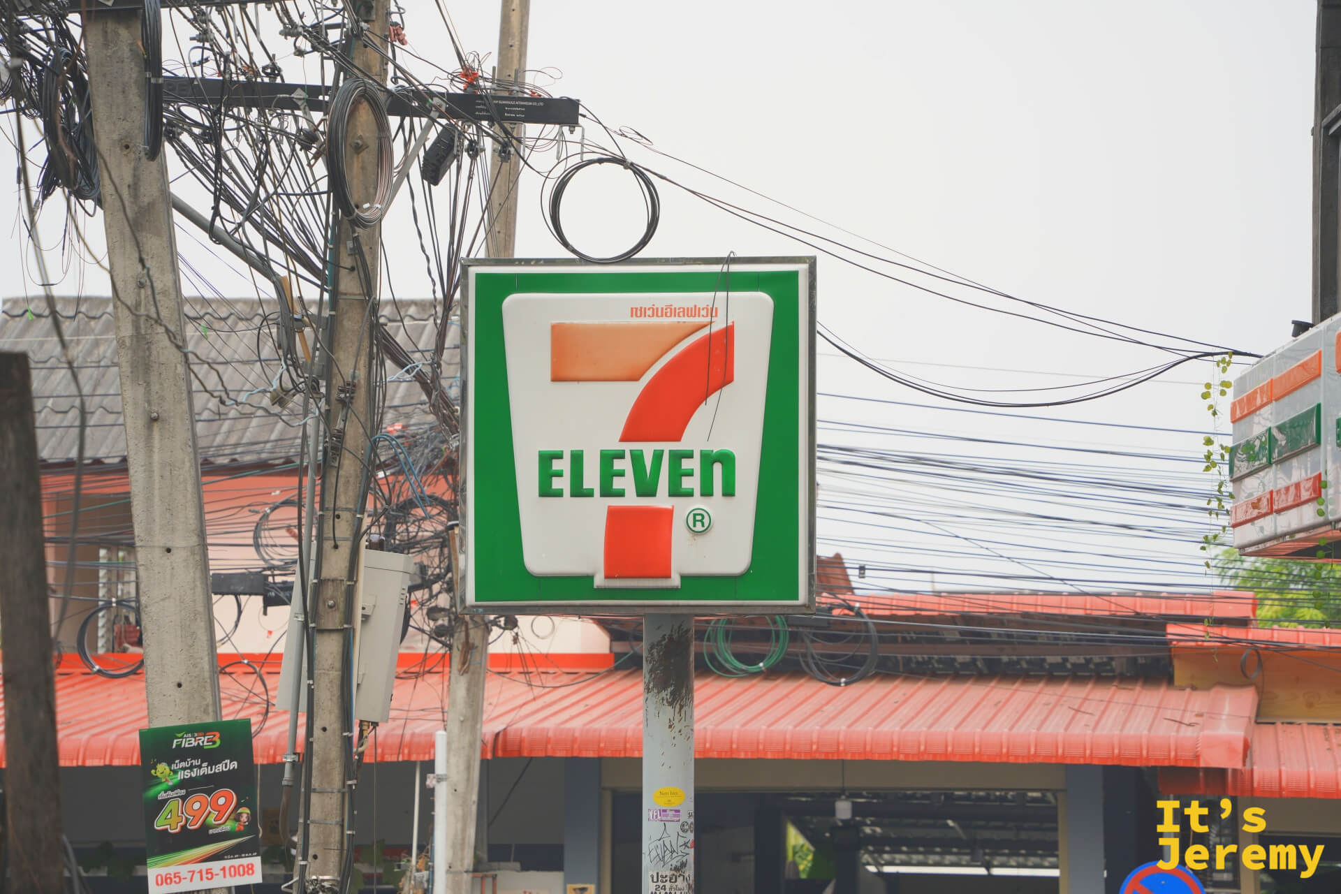 7-Eleven 招牌懸在街口,背後是糾纏交錯的電線與紅色屋頂