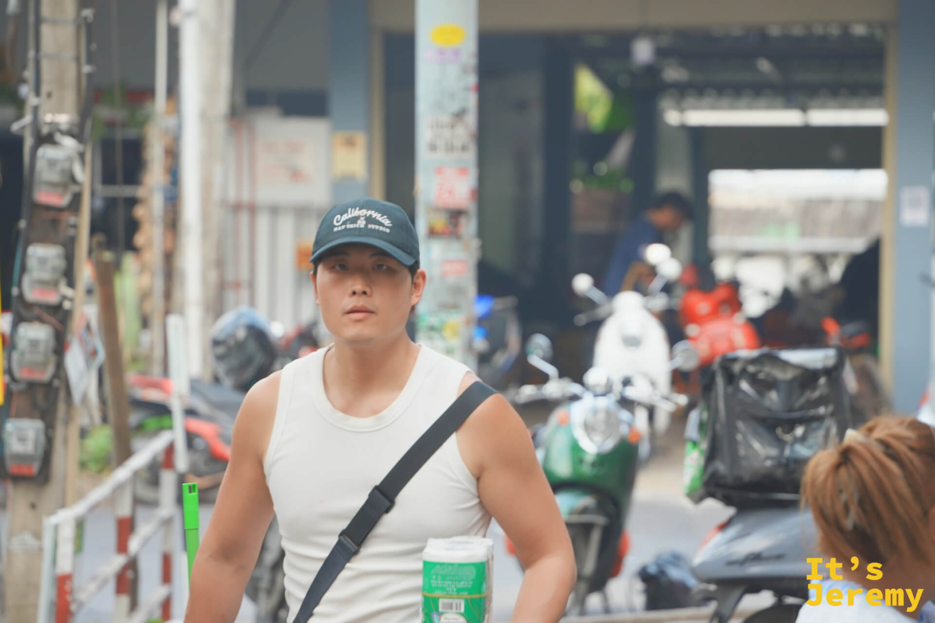 街邊一名戴棒球帽的男子抱著捲筒衛生紙走過,身後是停滿摩托車的騎樓