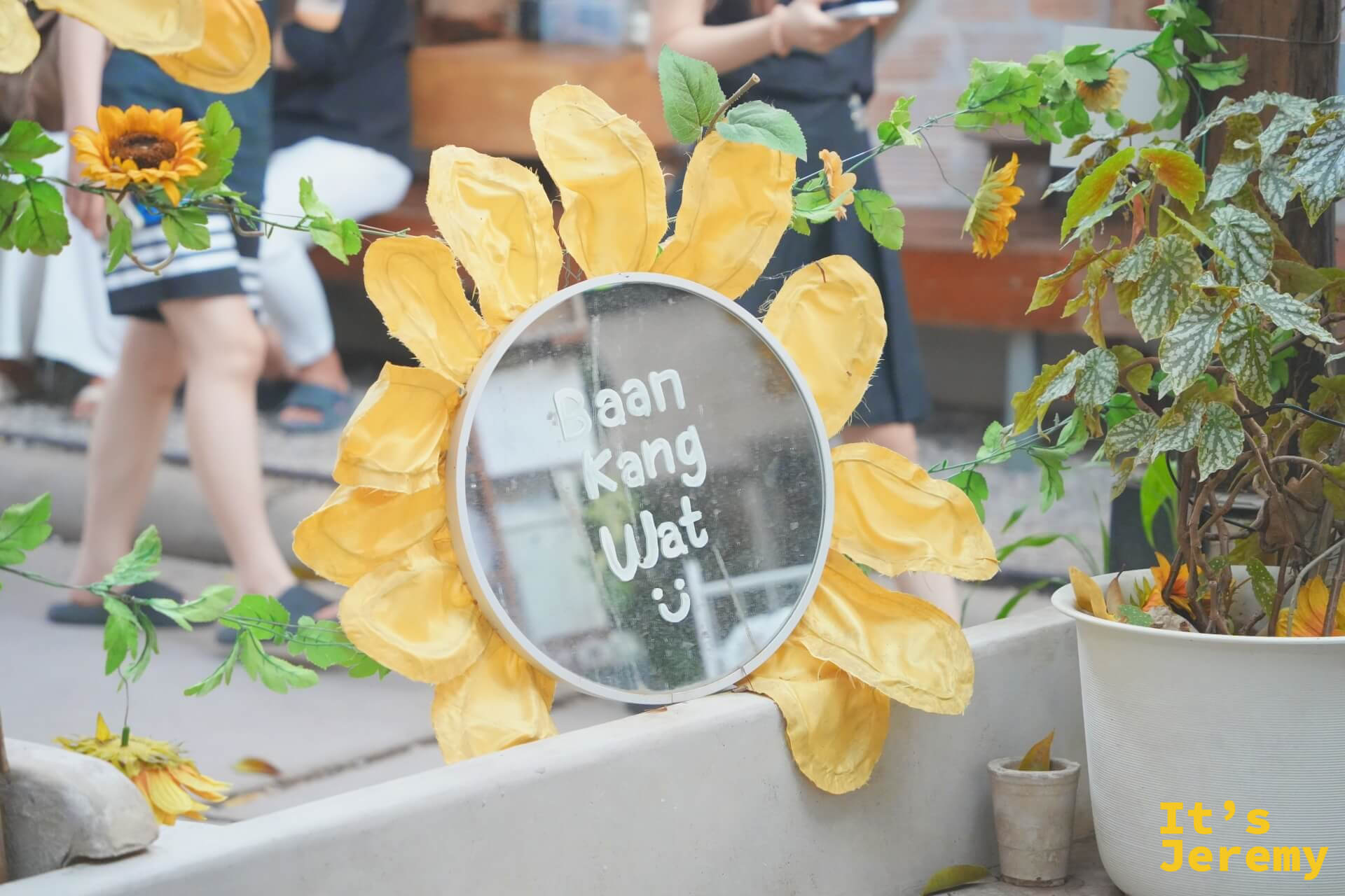 向日葵花框圓鏡,中央鏡面上寫著「Baan Kang Wat :)」,黃色布料花瓣圍繞,旁邊有盆栽和綠葉點綴