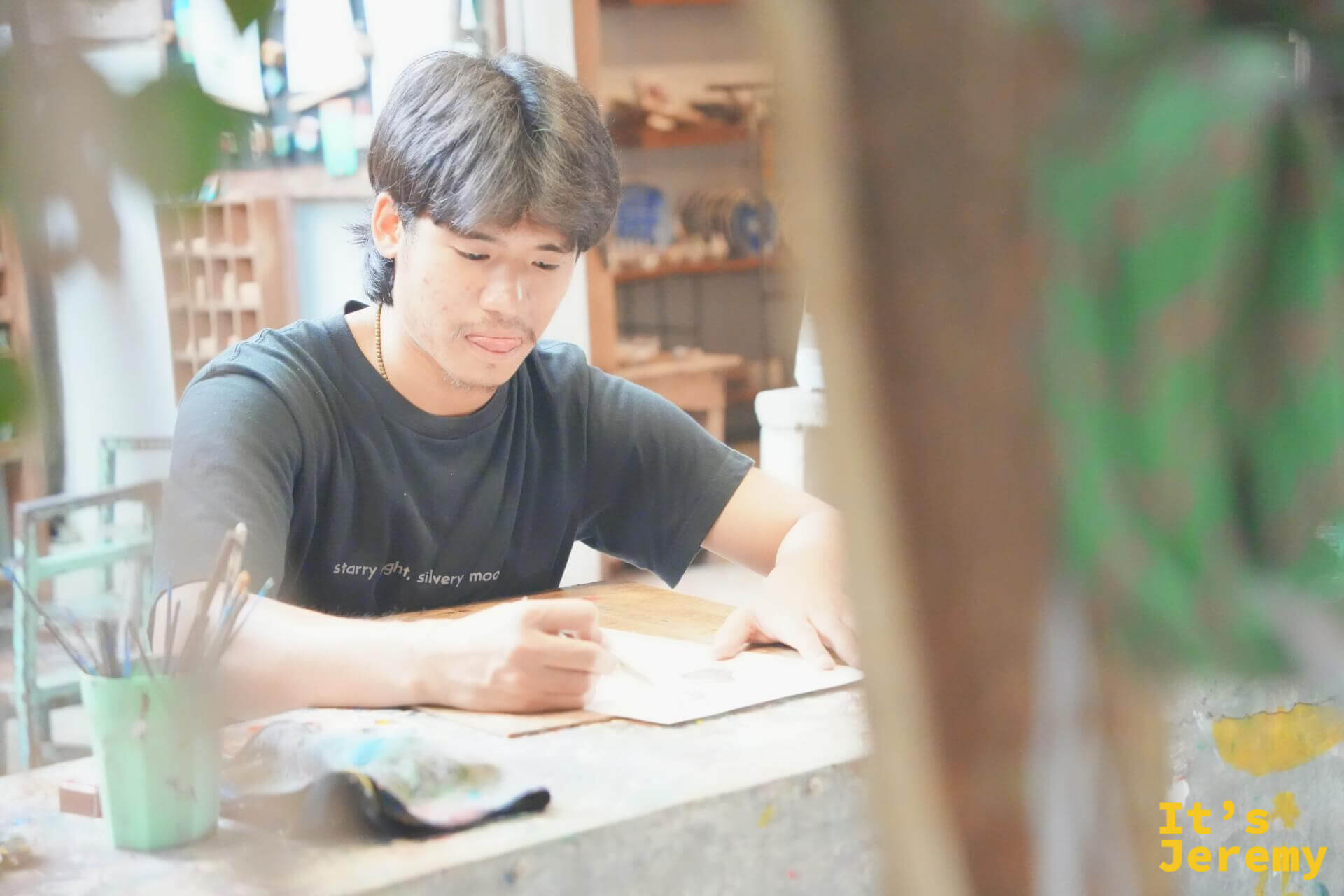 年輕男性在工作室內低頭創作,桌上有顏料調色盤和畫筆,後方是陶瓷作品展示架