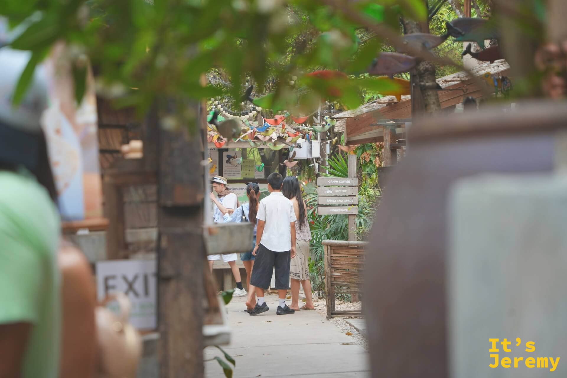 從植物葉叢間望向 Baan Kang Wat 廣場,幾個遊客背對,右側木製指示牌標著各店方向