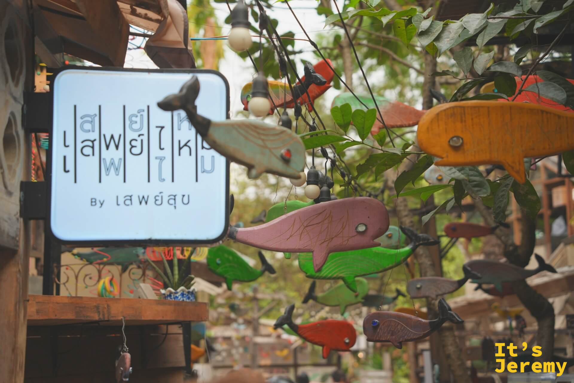 泰文店名招牌(เสพย์ไหม)旁,各色木雕魚從空中垂掛,粉色、綠色、棕色魚形在葉叢間搖動