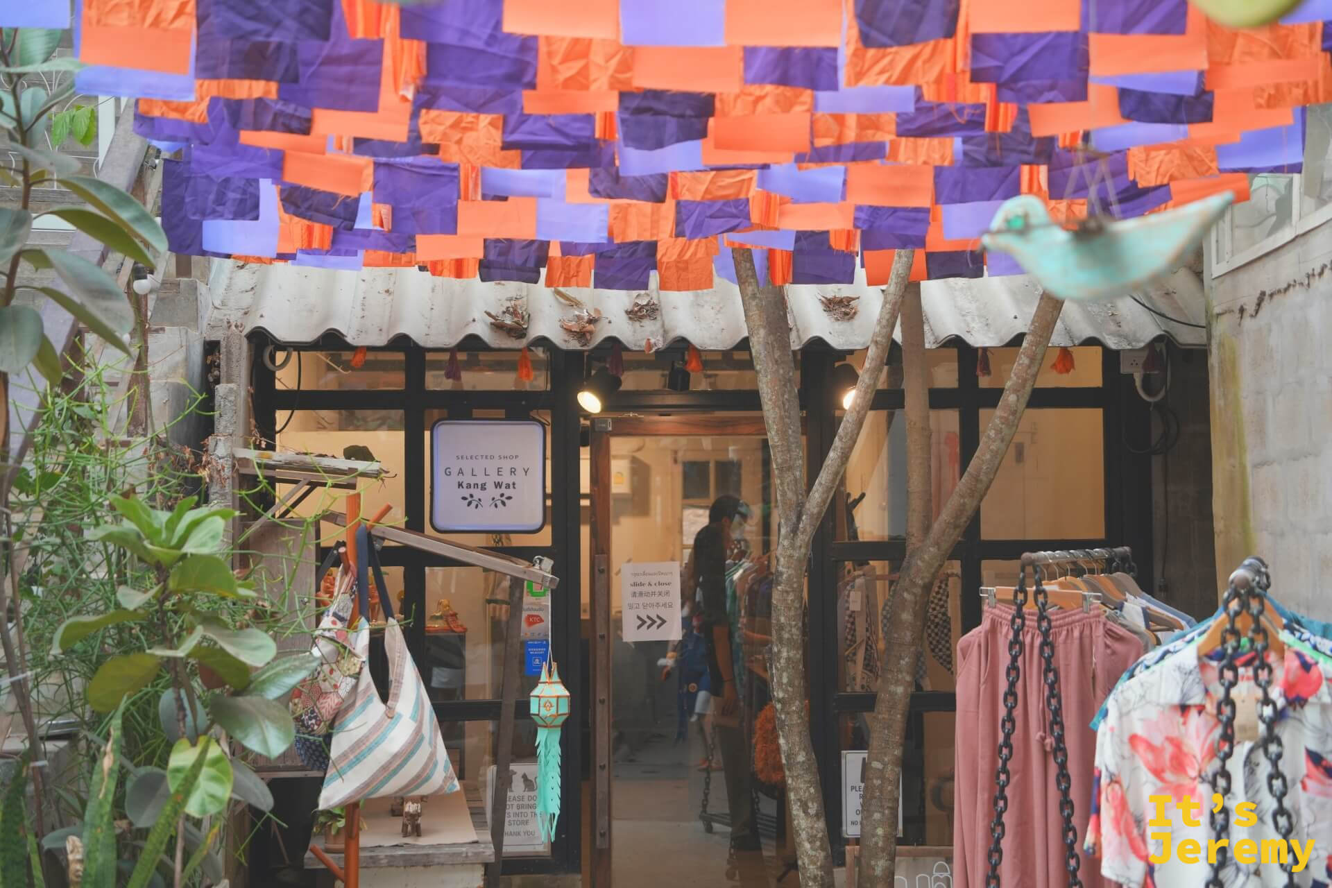 Gallery Kang Wat 選品店外觀,天花板懸掛橘紫布幔,店門口有衣架展示服裝,樹枝裝飾立在入口