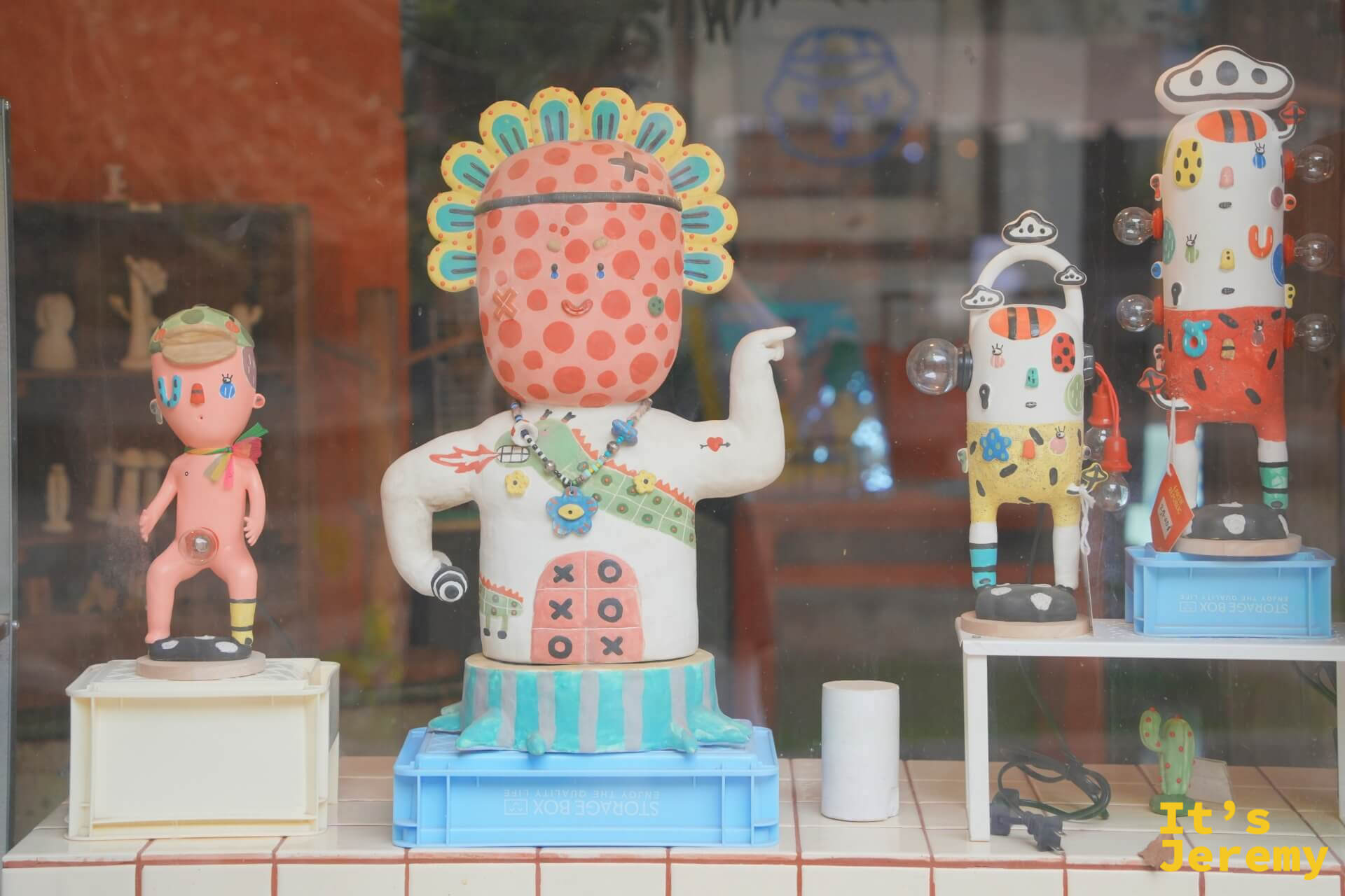 店家玻璃窗內展示多件彩色陶瓷藝術人偶,造型奇特,有太陽花頭冠、圓點斑紋和各種奇趣細節
