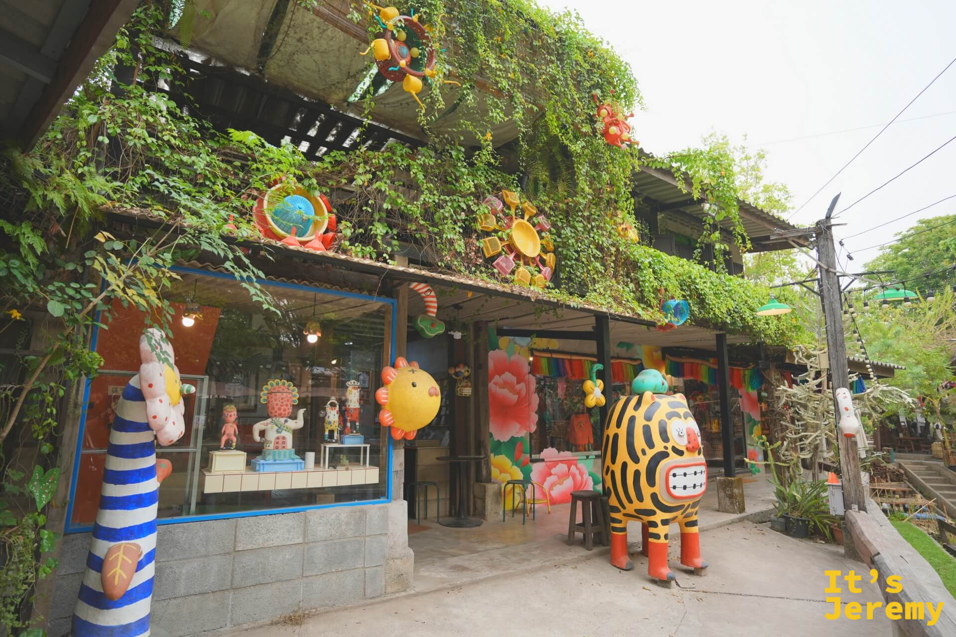 Baan Kang Wat 其中一棟建築外觀全景,爬牆植物茂盛,掛滿彩色藝術裝置,老虎吉祥物佇立門口