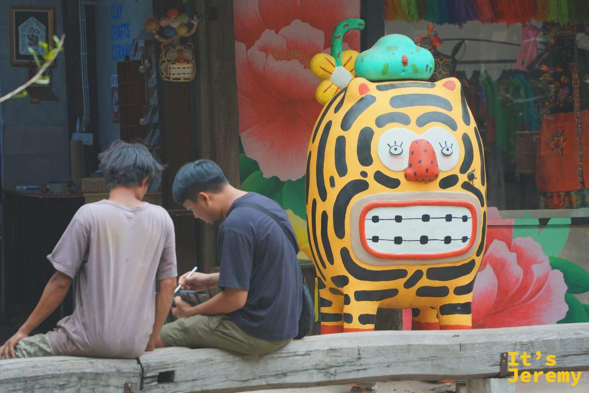 兩個男生坐在大型老虎吉祥物旁,神情悠閒,一人低頭看手機,旁邊可見彩色商品陳列