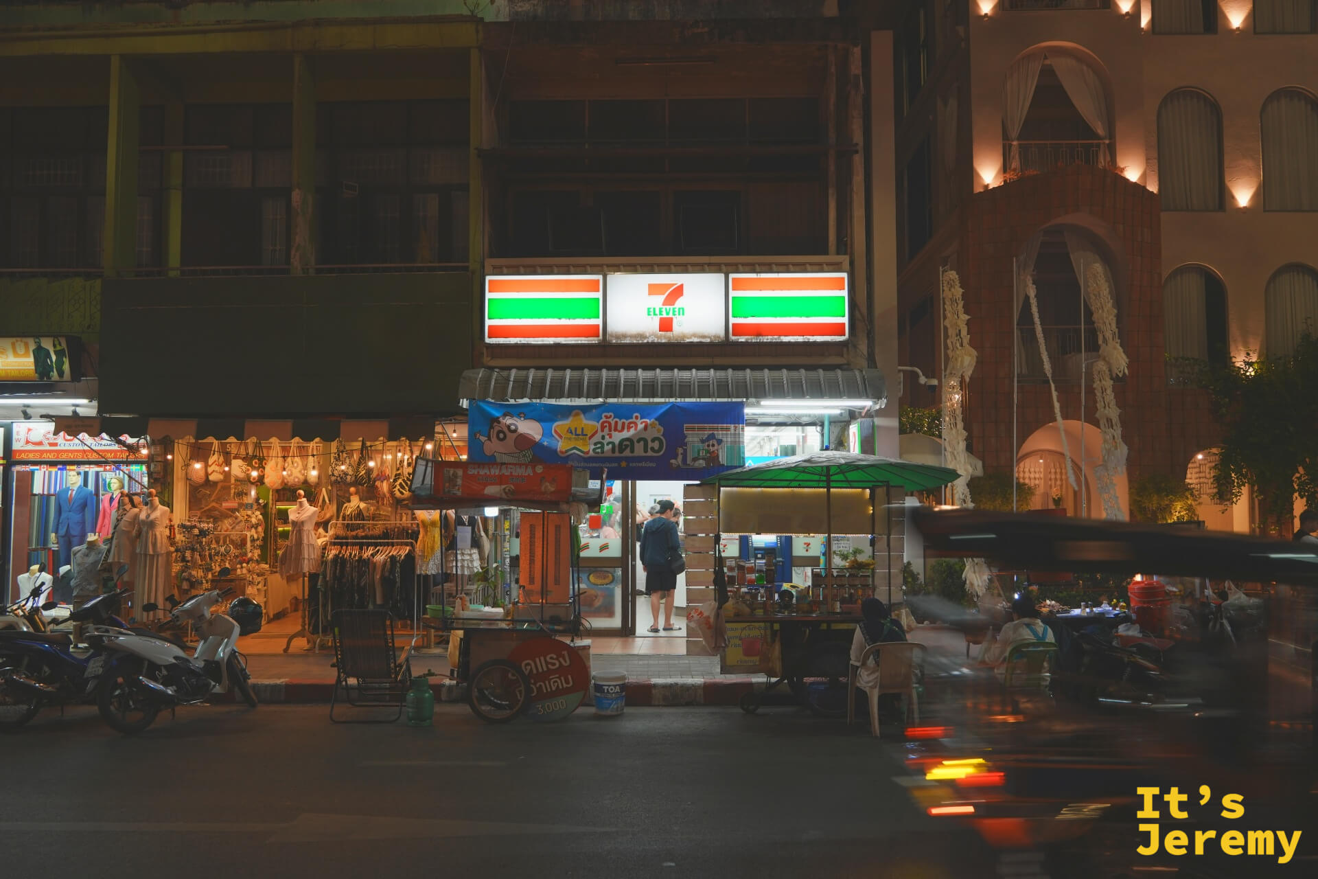夜裡的 7-Eleven 門口亮著燈,騎樓下有攤販與遮陽傘,店門口貼著蠟筆小新橫幅