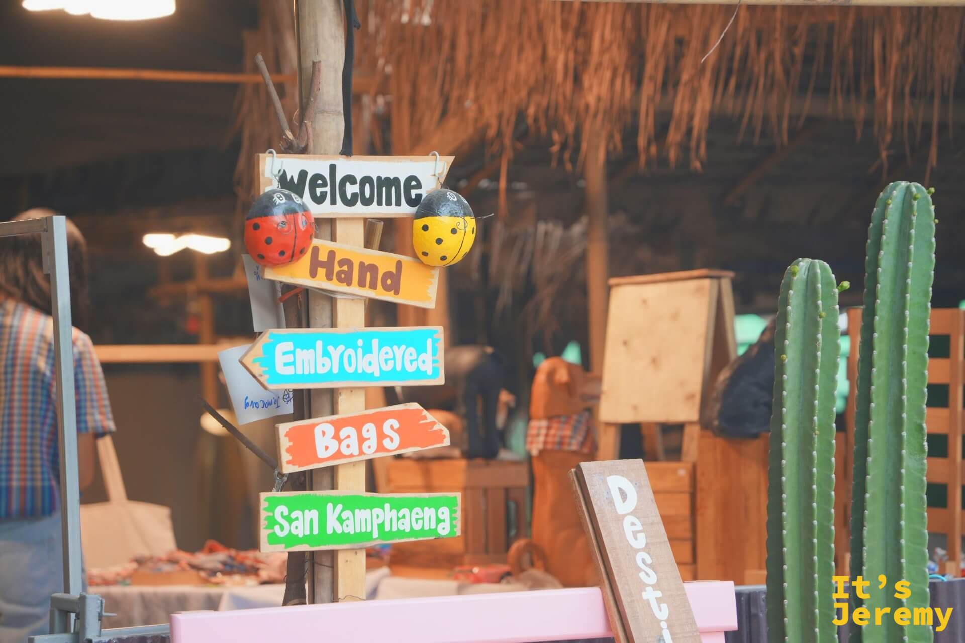 彩色手繪木牌指標,寫著「Welcome / Hand Embroidered Bags / San Kamphaeng」,旁邊立著兩株仙人掌