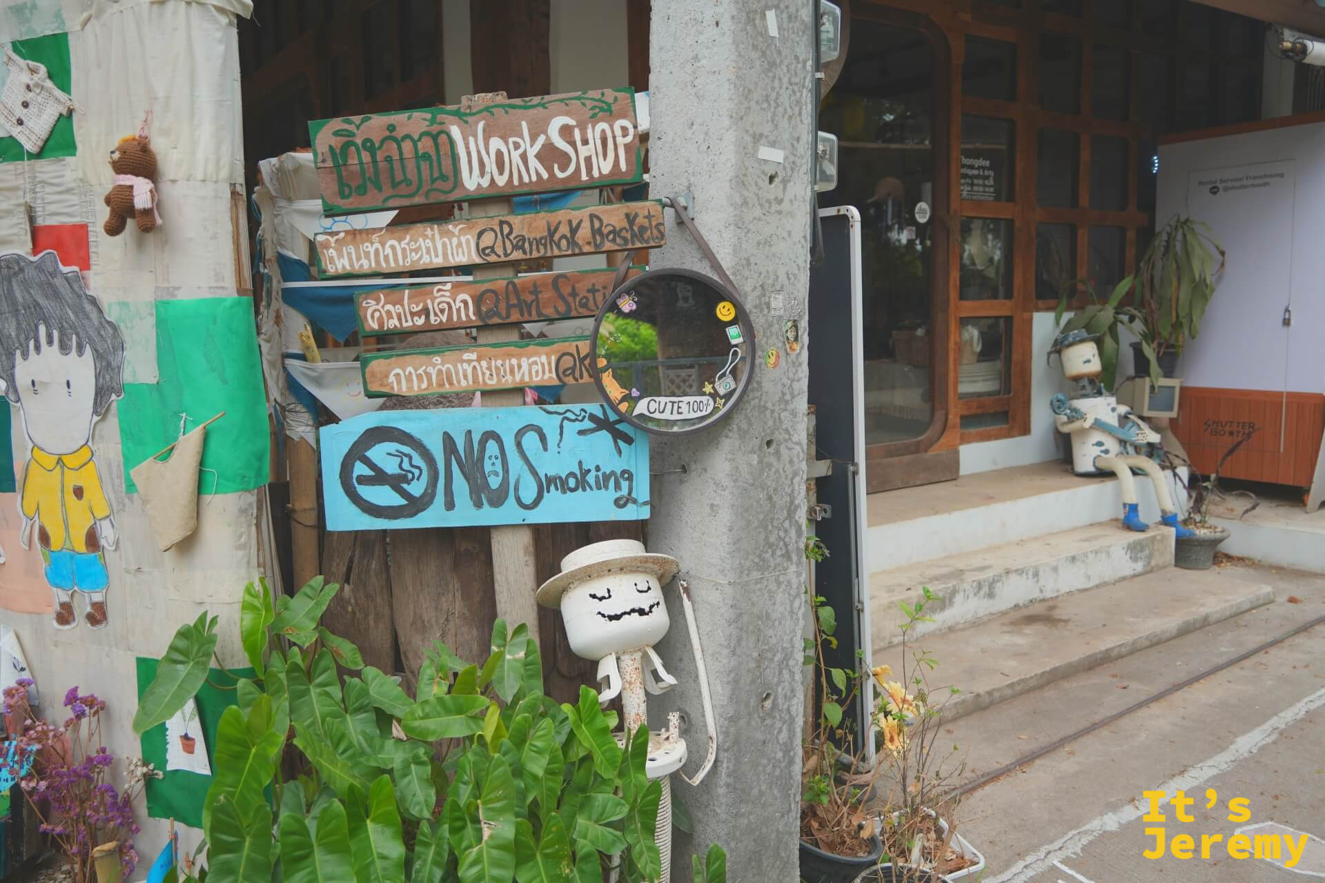 木牌指標柱上層層疊著 WORKSHOP、Bangkok Baskets、Art Studio 與 No Smoking 等手繪牌,下方站著一個戴帽子的棉花糖小人偶