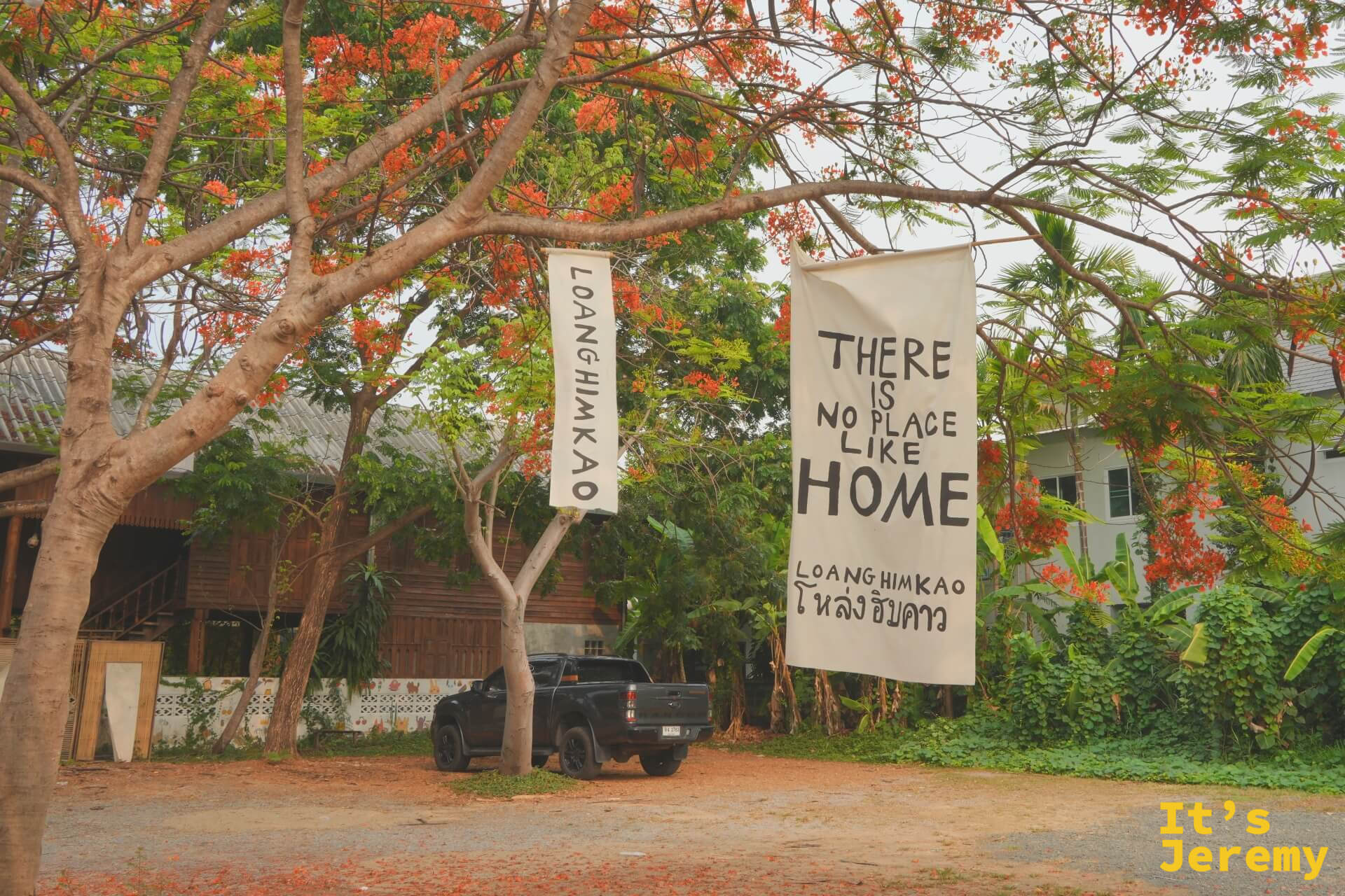 鳳凰花樹下掛著白色大布旗,寫著「THERE IS NO PLACE LIKE HOME — LOANGHIMKAO โหล่งฮิมคาว」