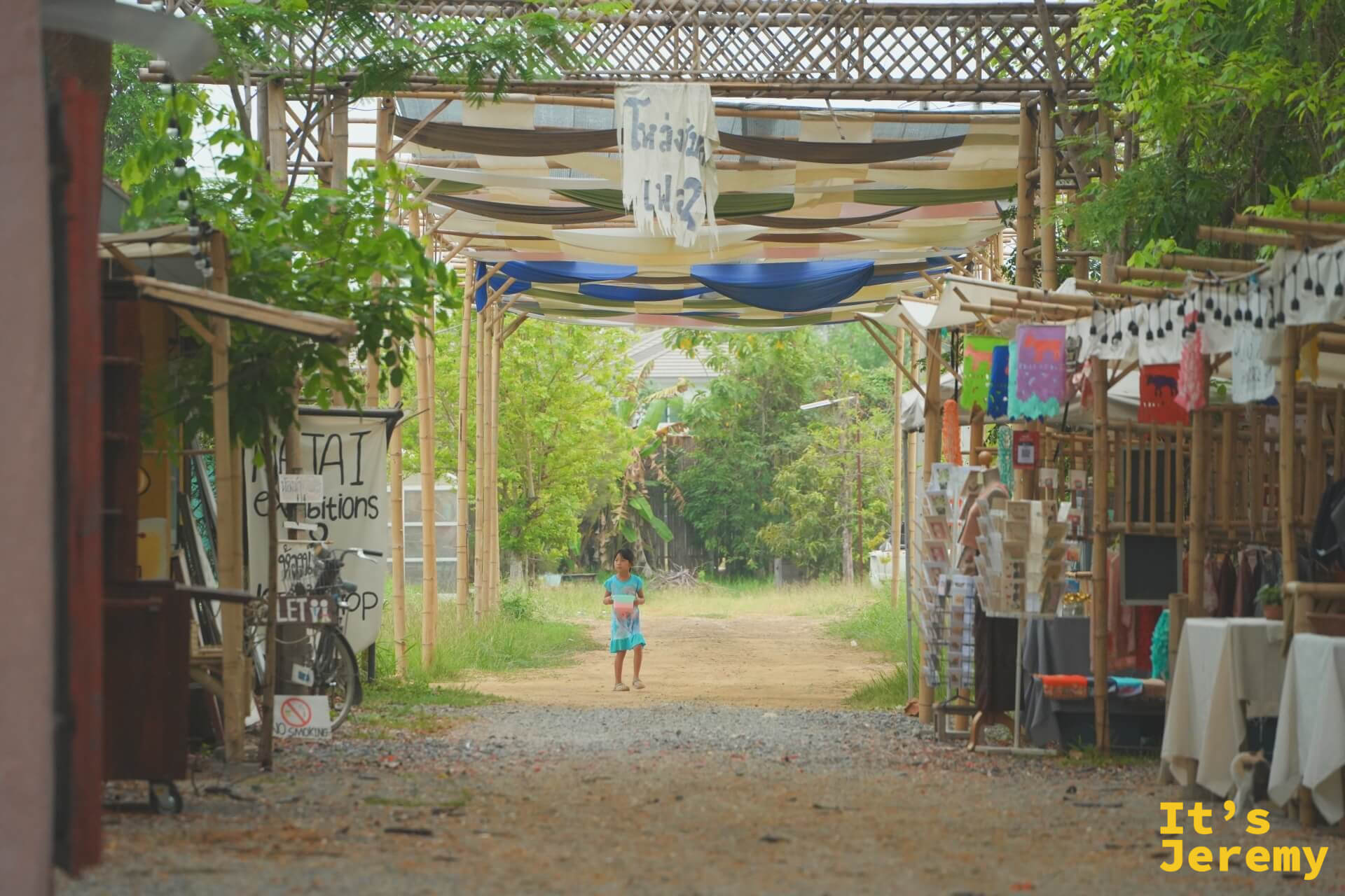 市集另一段巷道的布條天棚下,一個小孩走在碎石路中央,兩側攤位正在準備