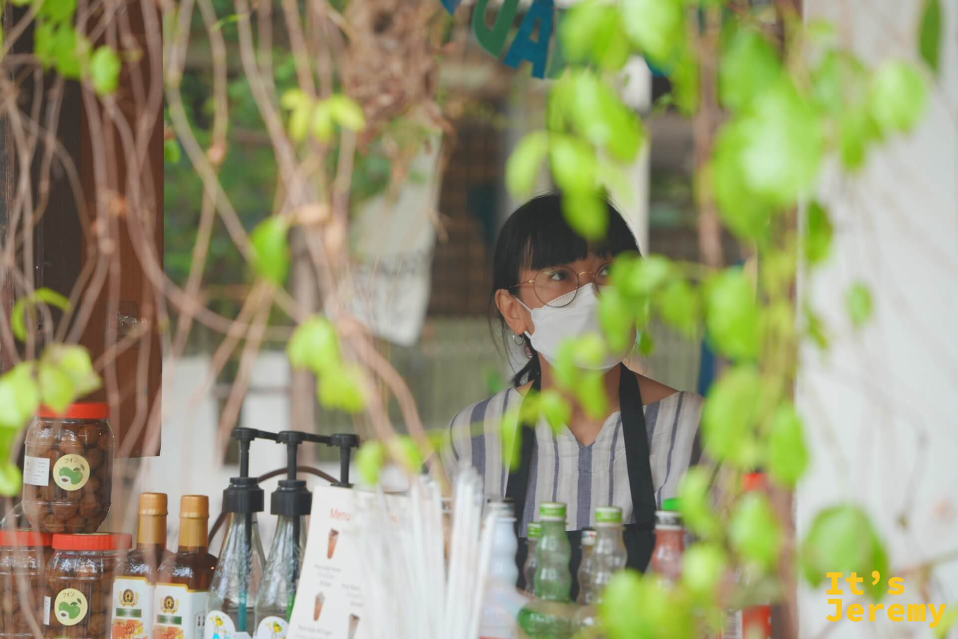 透過藤蔓縫隙看 EDUMade 的老闆娘,戴著眼鏡與口罩站在攤位後方