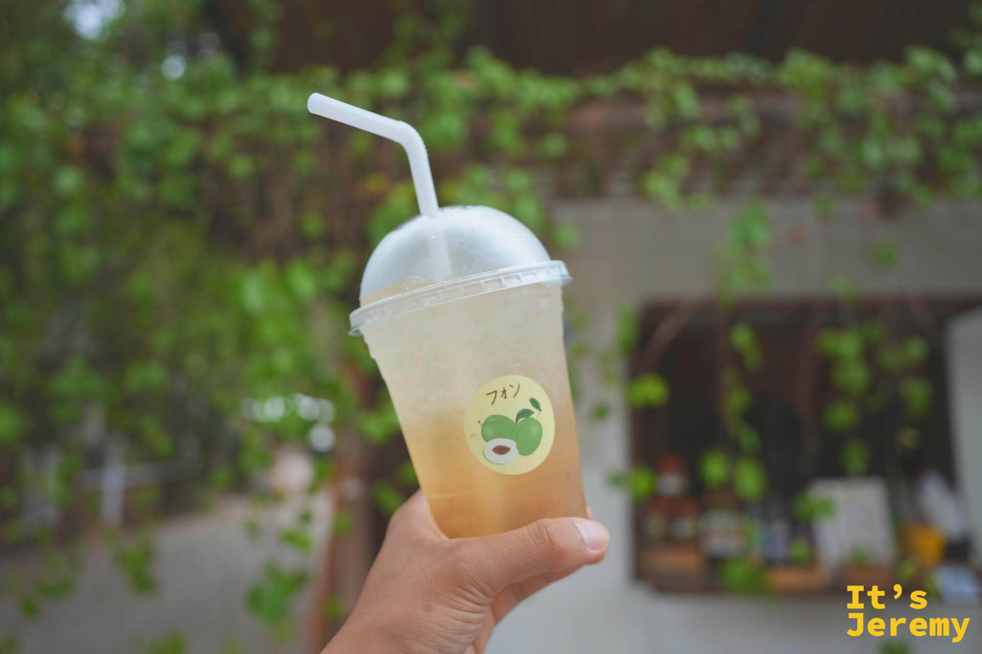 手拿一杯 EDUMade 的梅子蘇打,杯身貼著綠梅貼紙,背景是藤蔓垂落的店面