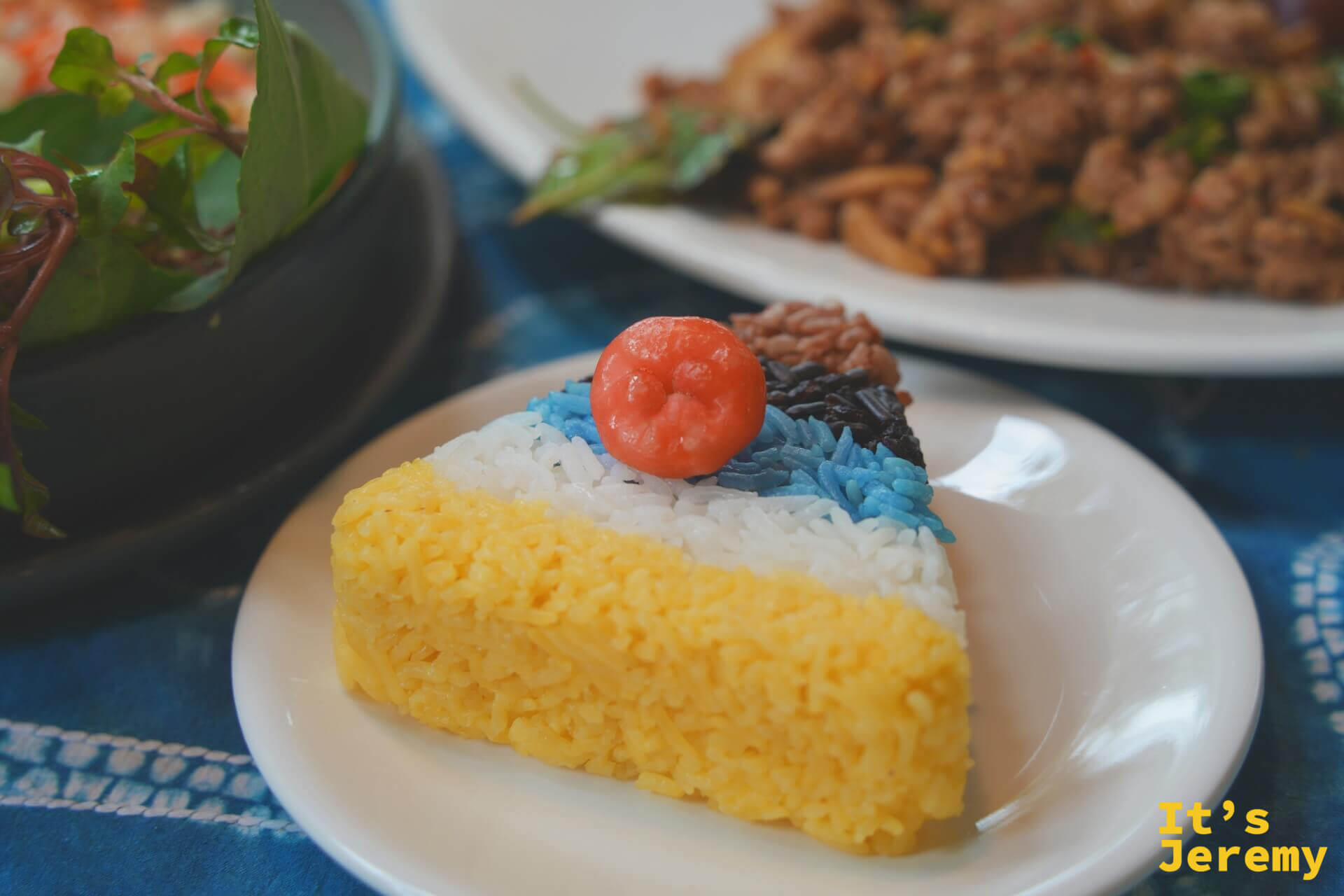 五色米飯特寫，三角形如蛋糕切片，底層薑黃色、中層白色、頂層藍紫黑三色，頂端放著一顆紅色小球