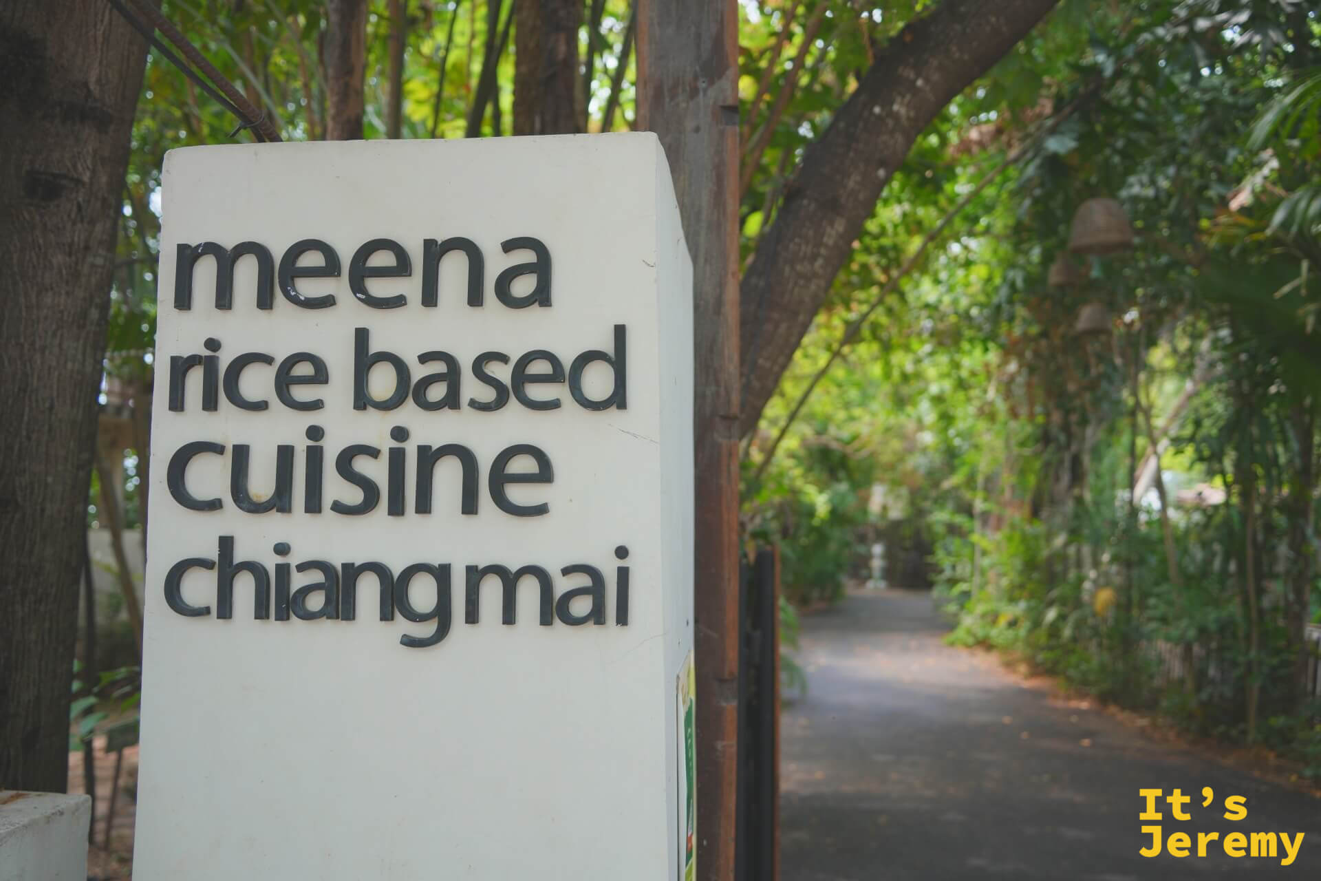 白色方柱招牌寫著 meena rice based cuisine chiangmai，後方是綠蔭步道與掛著銅鈴的樹
