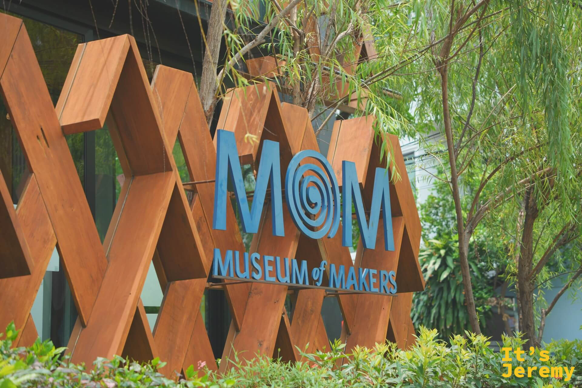 木格柵上的藍色 MOM(Museum of Makers)立體字標誌,周圍被樹葉環繞