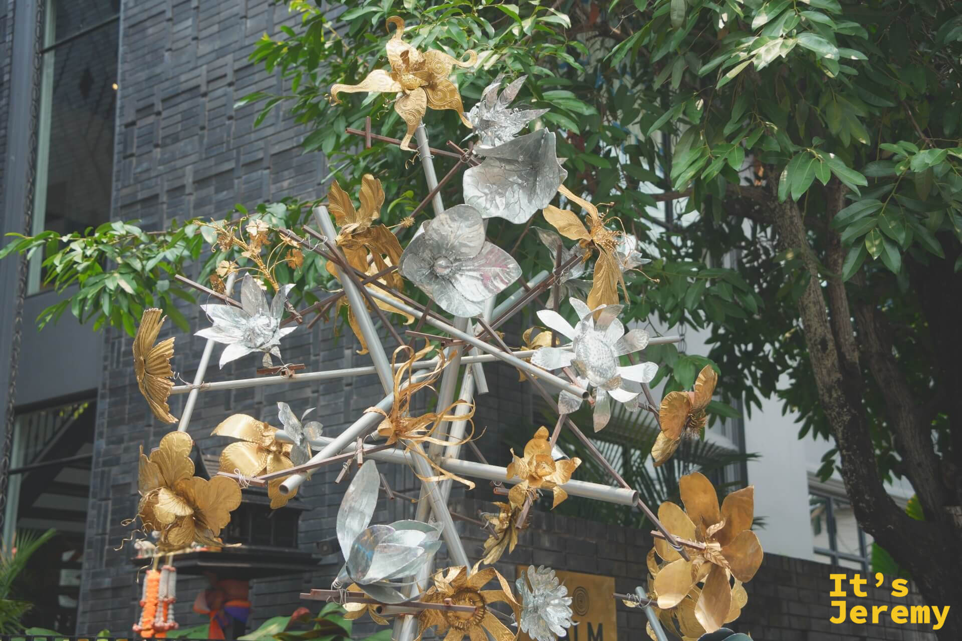 銀色與金色金屬花朵組成的雕塑,立在灰磚建築前的綠樹之間