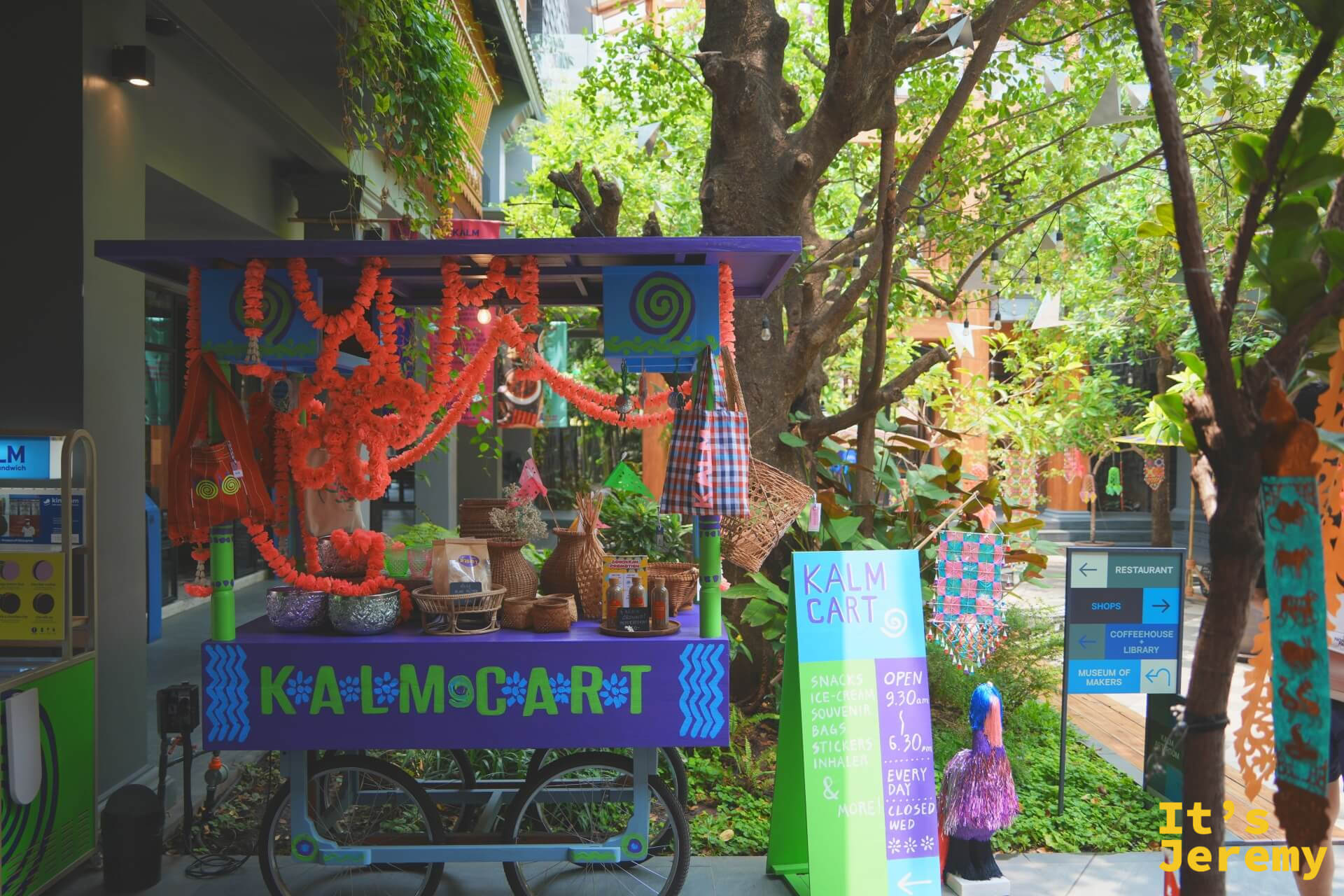 紫色的 KALM CART 小攤車,掛滿橘色花環與藤編籃,停在中庭樹下