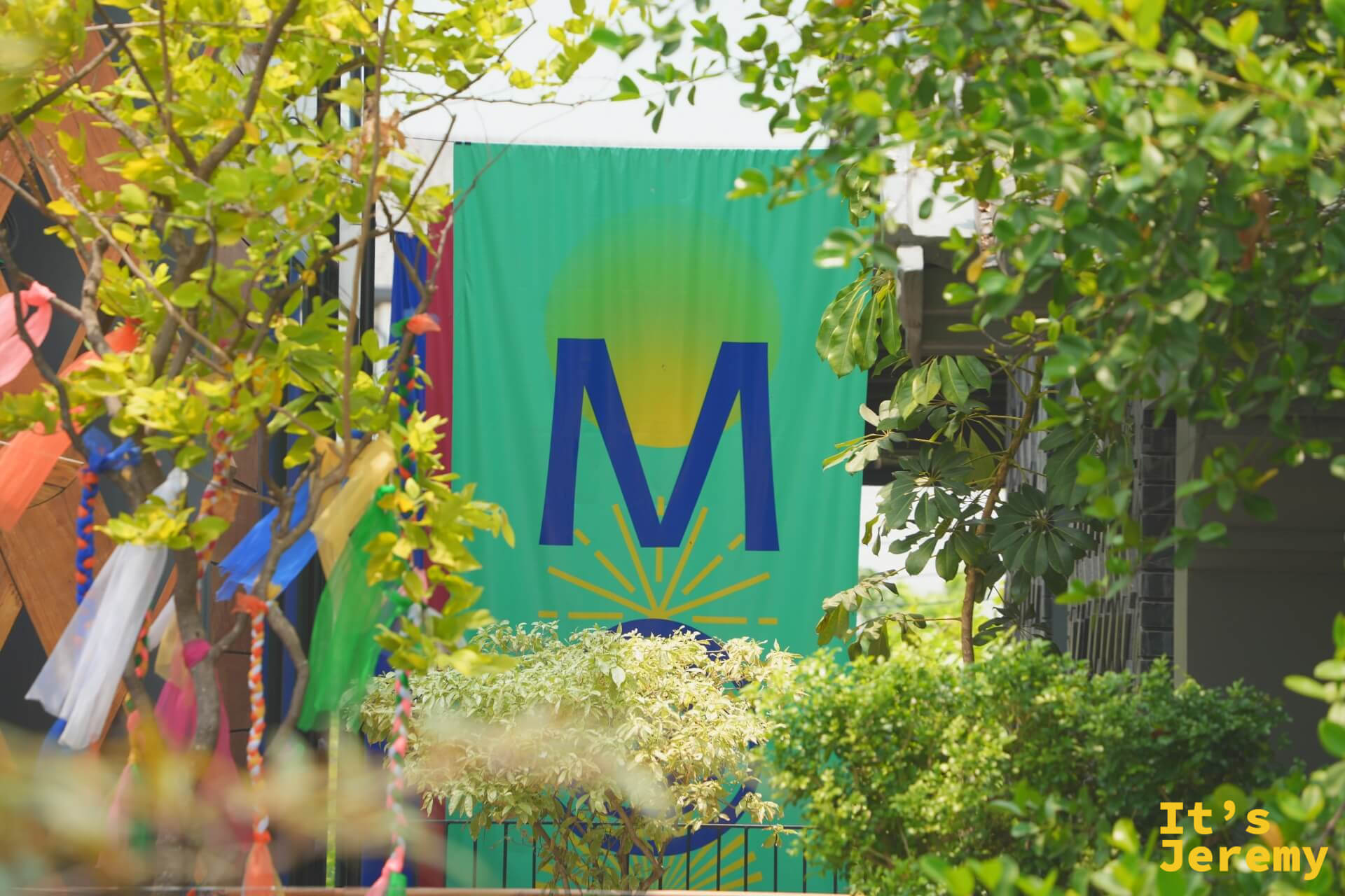 樹叢間露出 MOM 的綠底藍字旗幟,M 字下方是一輪放射狀的日出圖案