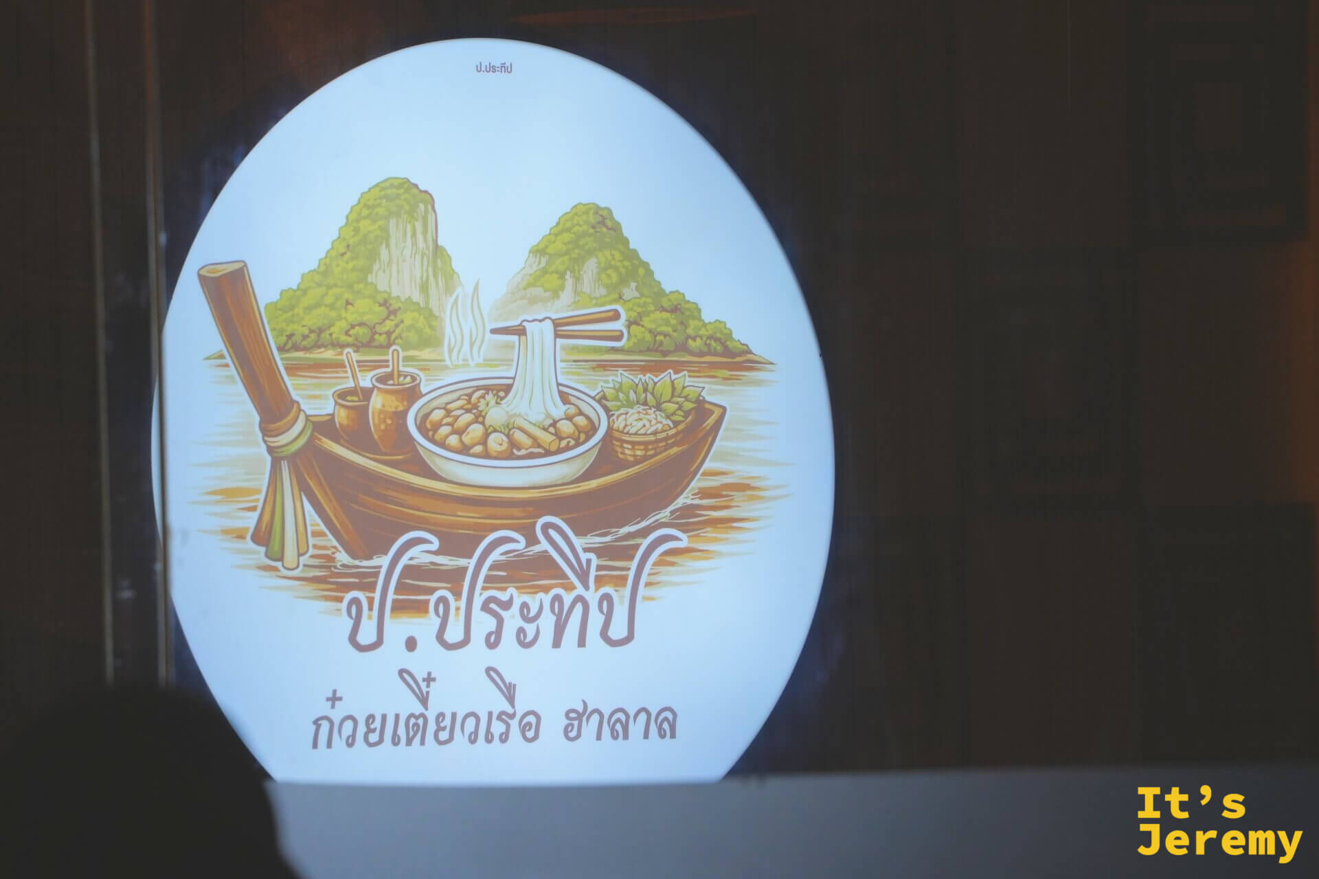 圓形燈箱招牌上畫著一艘長尾船，船上載著冒煙的船麵與調味罐，背景是喀比的石灰岩山與水面