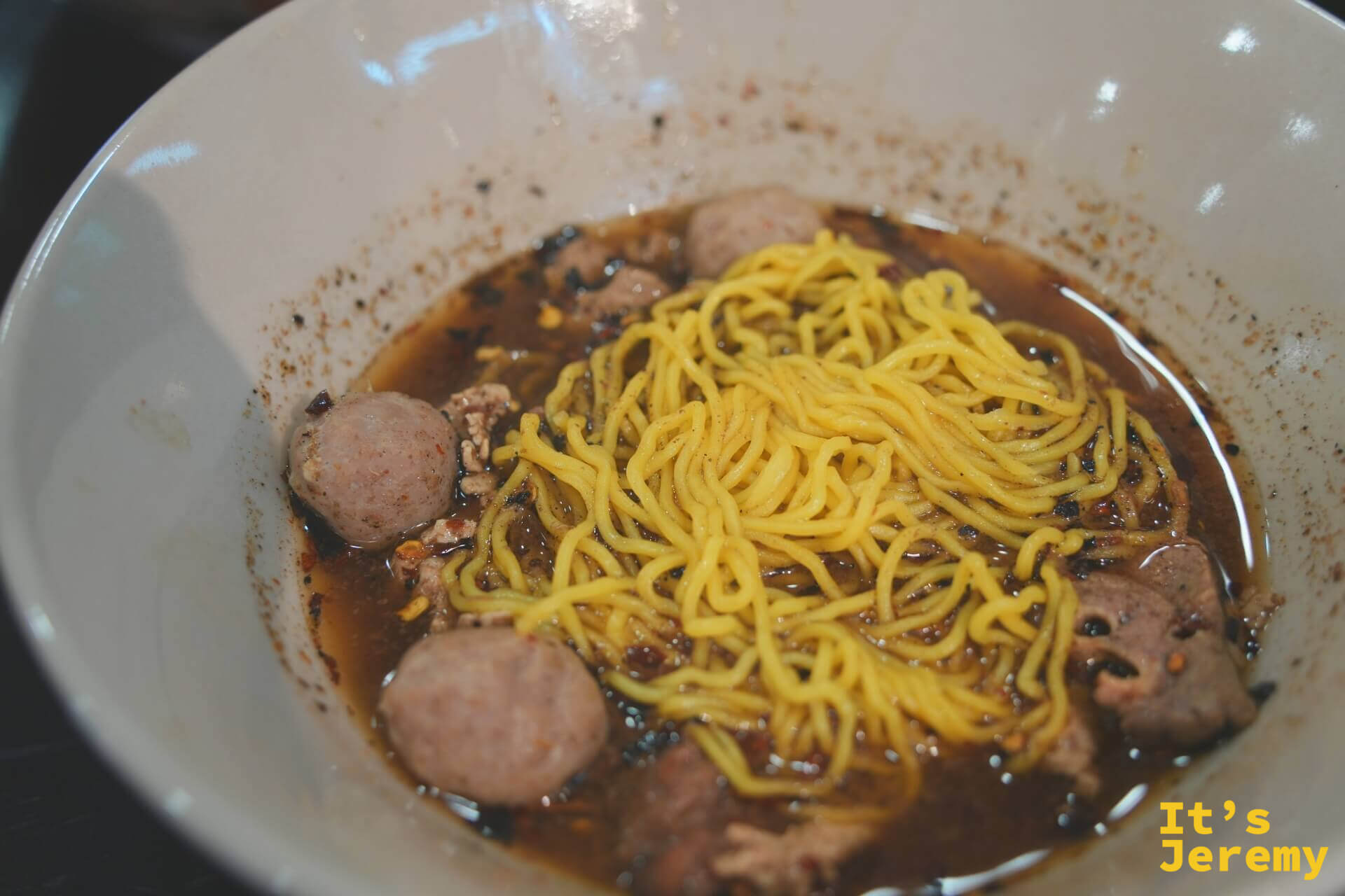 白碗裡的牛肉船麵，黃色雞蛋麵浸在深色辣湯中，湯裡有多顆牛肉丸與肉末，碗緣沾著辣椒碎