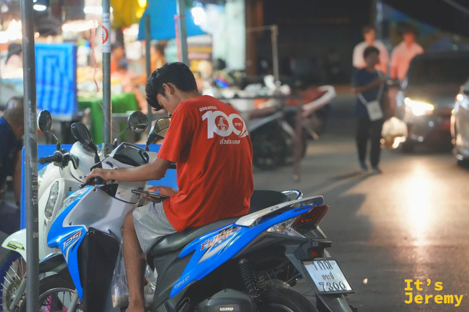 穿紅色上衣的少年跨坐在藍白色機車上低頭滑手機，背景是燈火通明的夜市攤位與來往人群