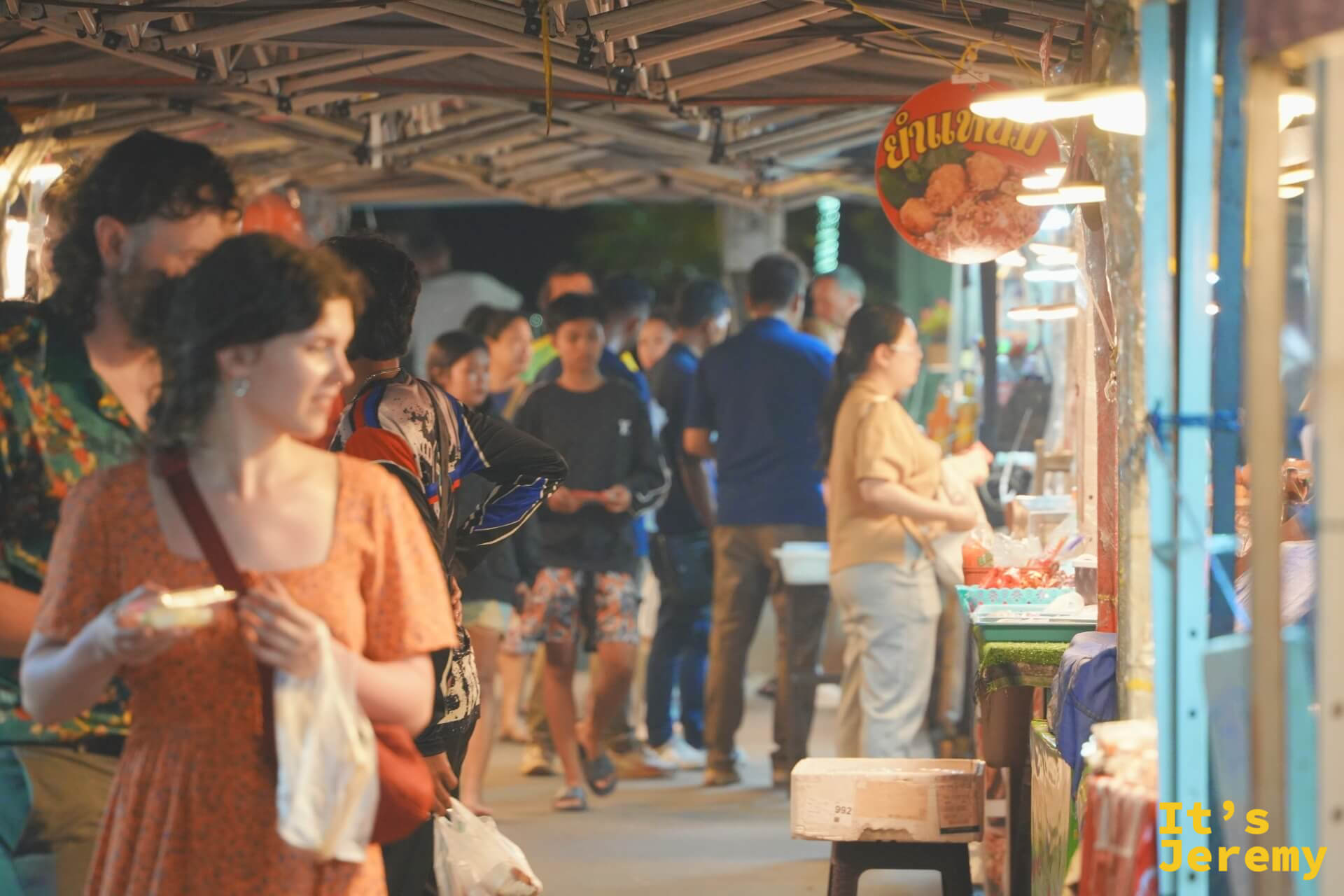 夜市帳棚下的走道擠滿人潮，前景一對外國旅人正端著小吃邊走邊吃，右側攤位掛著泰文圓形招牌
