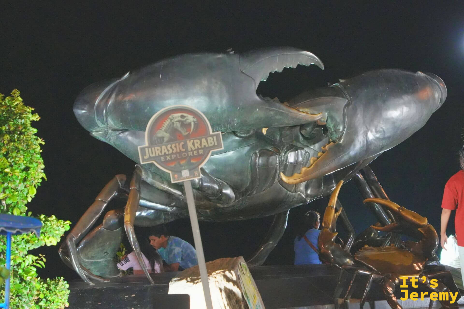 打燈的巨大黑蟹雕像在夜空下展開雙螯，前方立著 Jurassic Krabi Explorer 看板，雕像旁還有一隻金色螃蟹雕塑