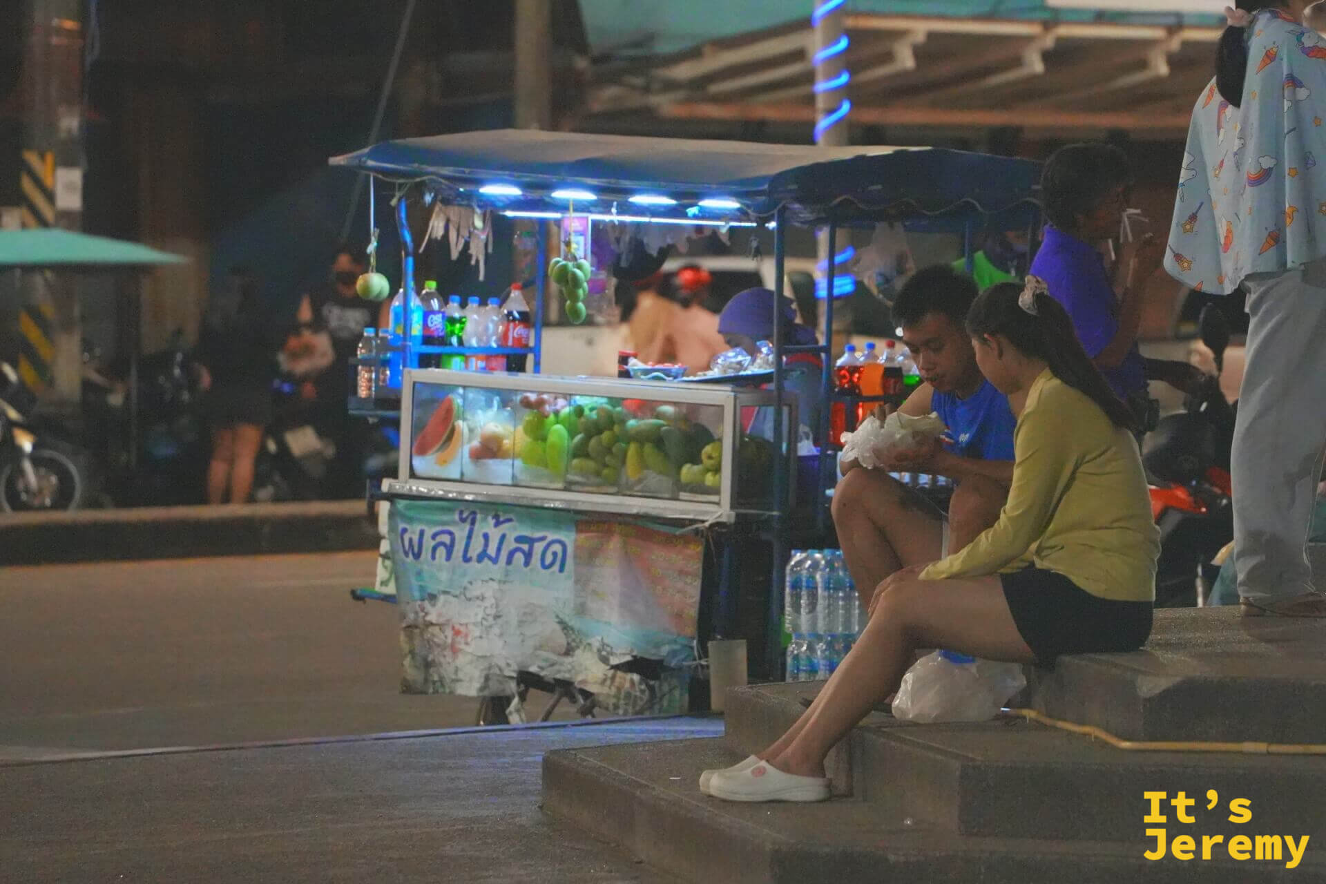 寫著泰文「新鮮水果」布條的水果攤前，一對年輕男女坐在階梯上分食袋裝水果