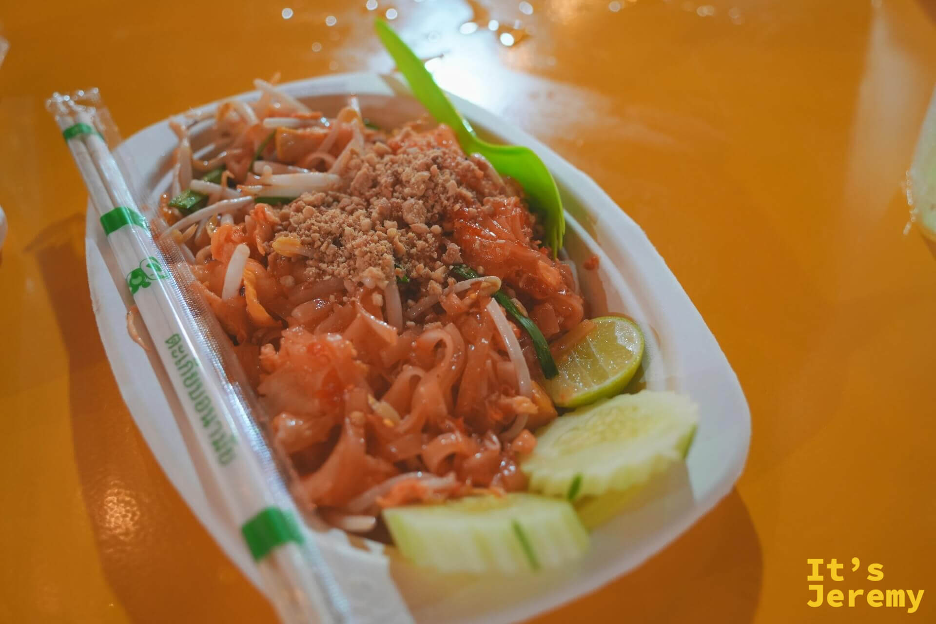 紙盤裝的泰式炒河粉，上面撒滿花生粉，旁邊配著黃瓜切片與一角萊姆，附綠色塑膠叉