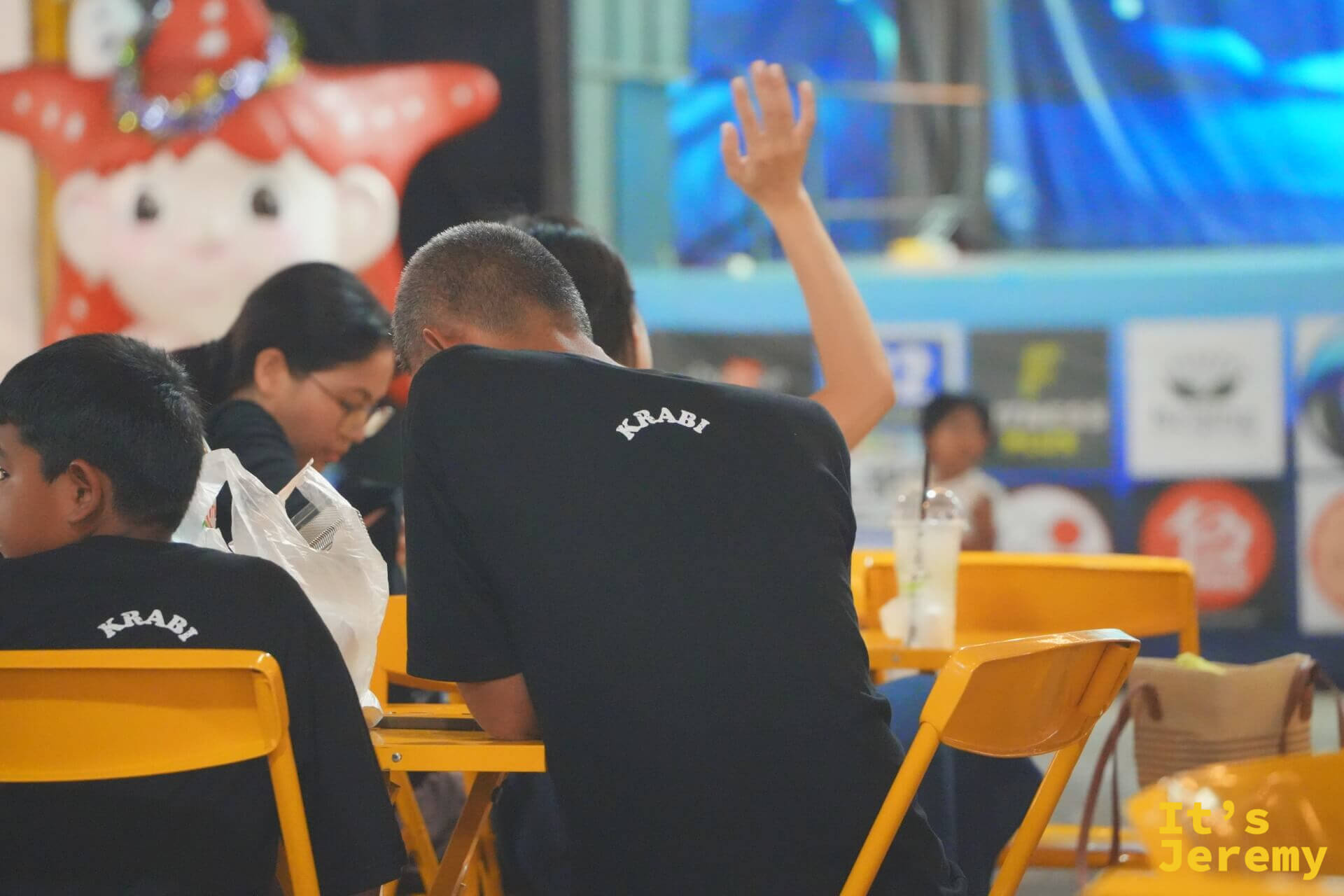 用餐區黃色桌椅旁坐著數名穿黑色 KRABI 字樣 T 恤的遊客，背景有一隻紅色海星造型的人偶裝飾