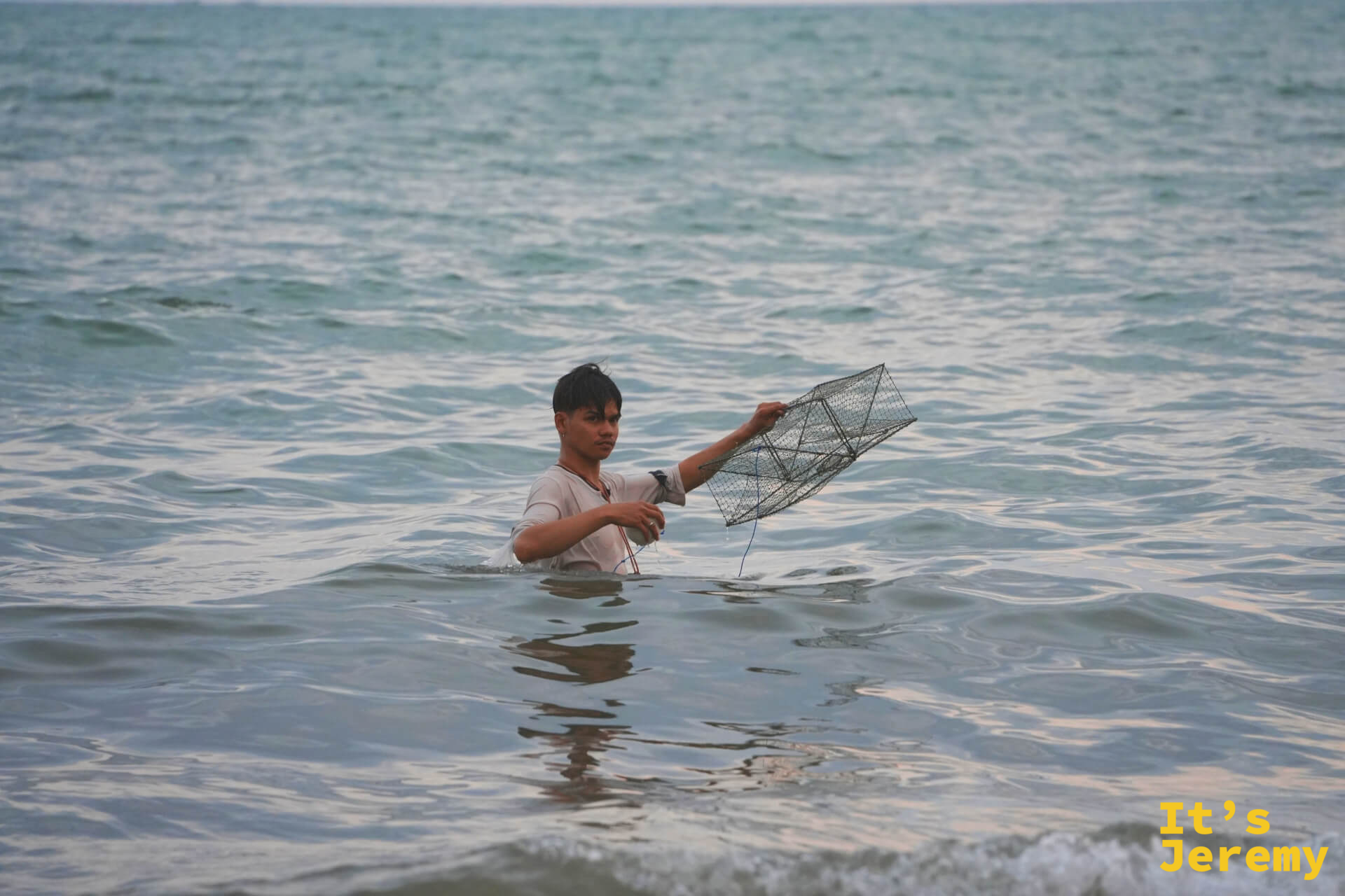 在地漁民站在齊腰深的海水中，手持方形金屬捕魚籠，神情專注