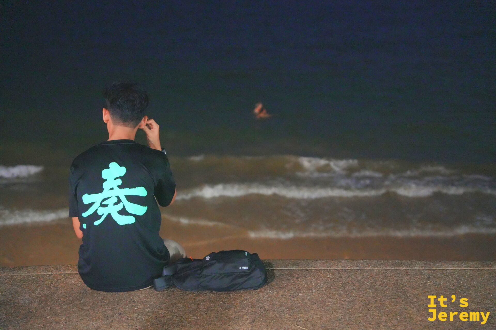 入夜後一名旅人背對鏡頭坐在低矮圍牆邊，望向深黑色海面，遠處有人在夜間游泳