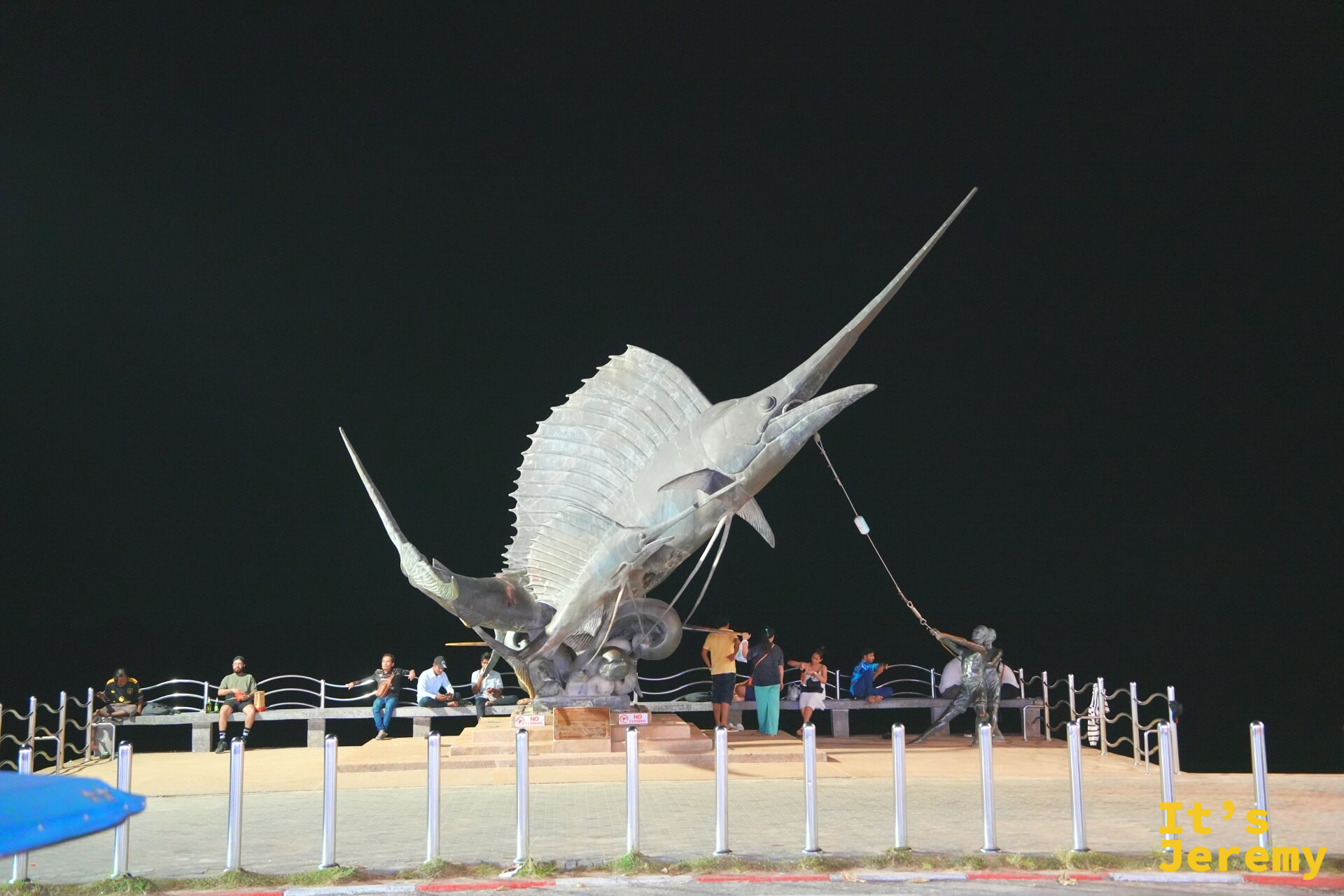 奧南廣場的大型銅色旗魚雕塑，夜燈下幾個旅人坐在底座休息