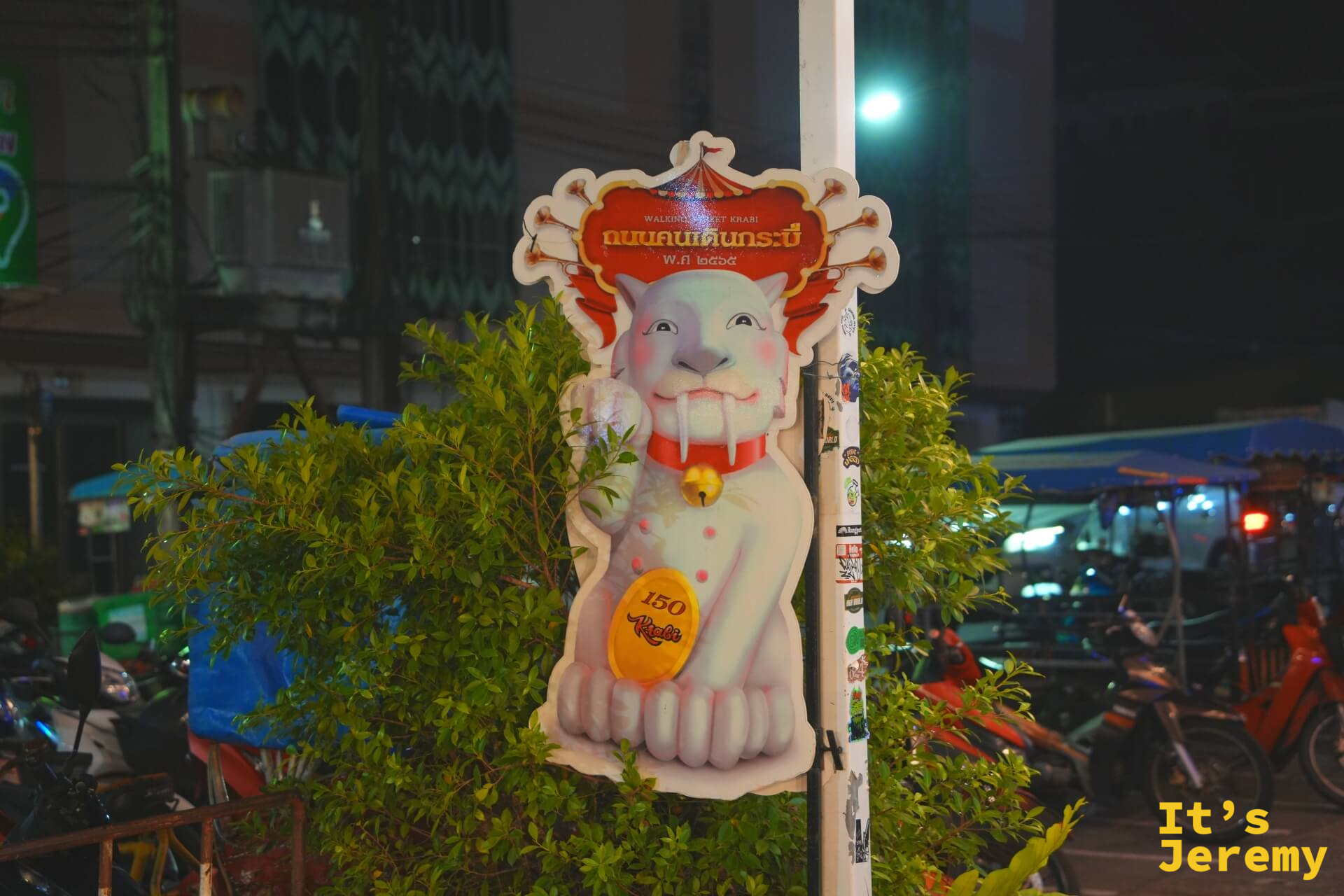 喀比鎮夜晚路口的 Walking Street Krabi 指示立牌,白色惡魔貓咪圖案立在植物叢前,背景是摩托車與夜市燈光