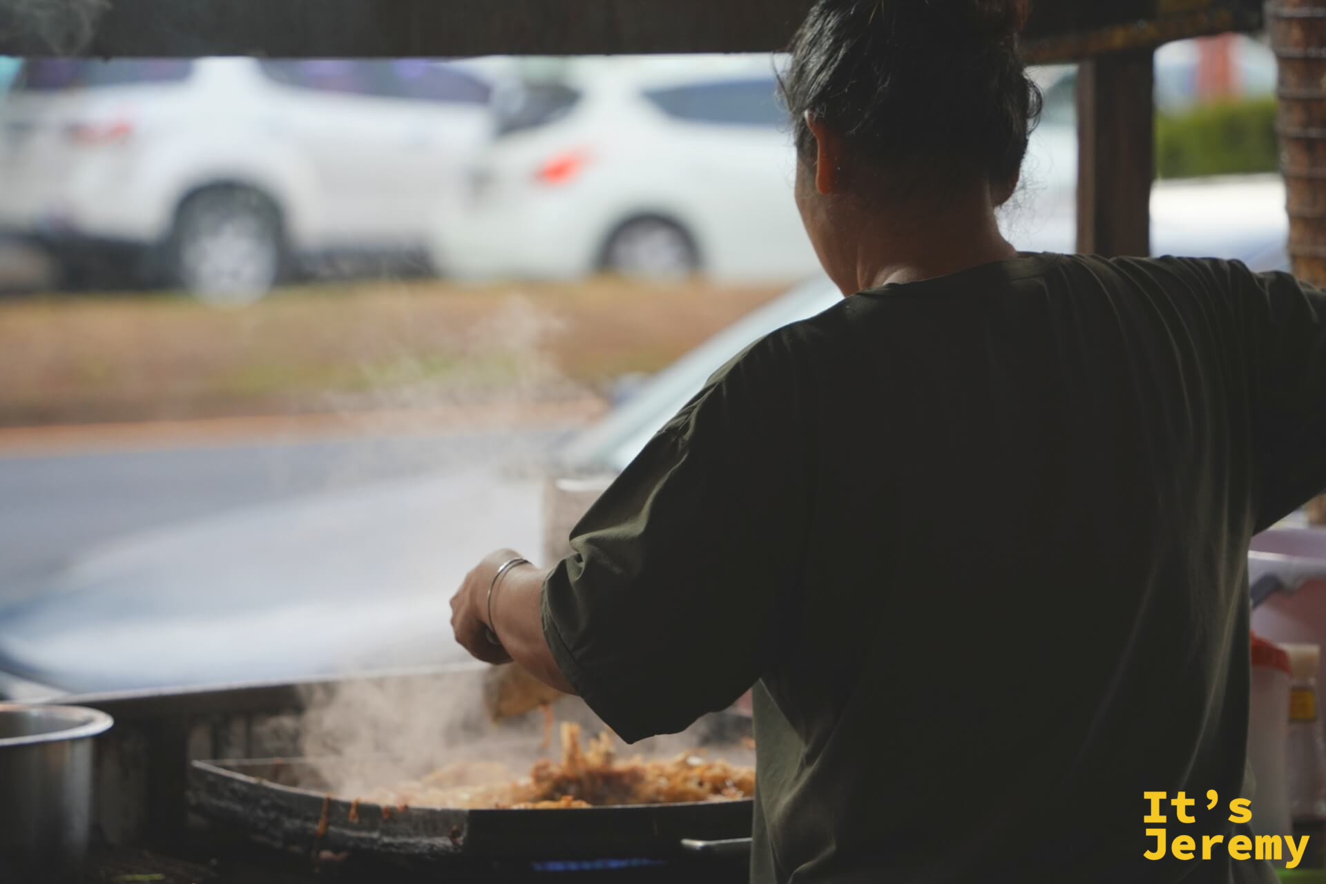 師傅背對鏡頭，全神貫注地在大圓鍋裡翻炒，白色油煙往棚外飄散
