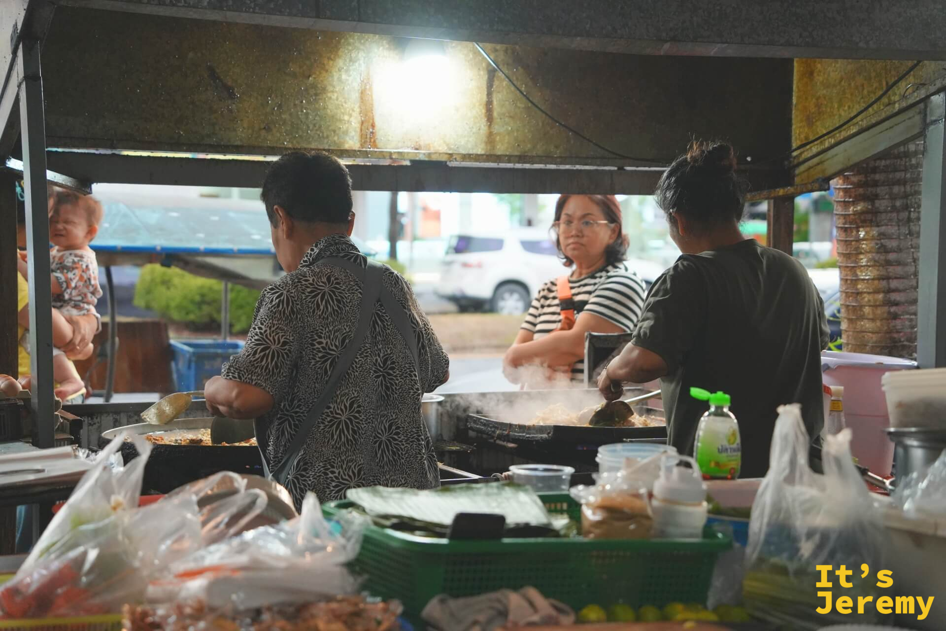 全家人一起工作的場景：一位大姐正在炒菜，背景有一名抱著嬰兒的女士和一名男性助手，棚外是街道