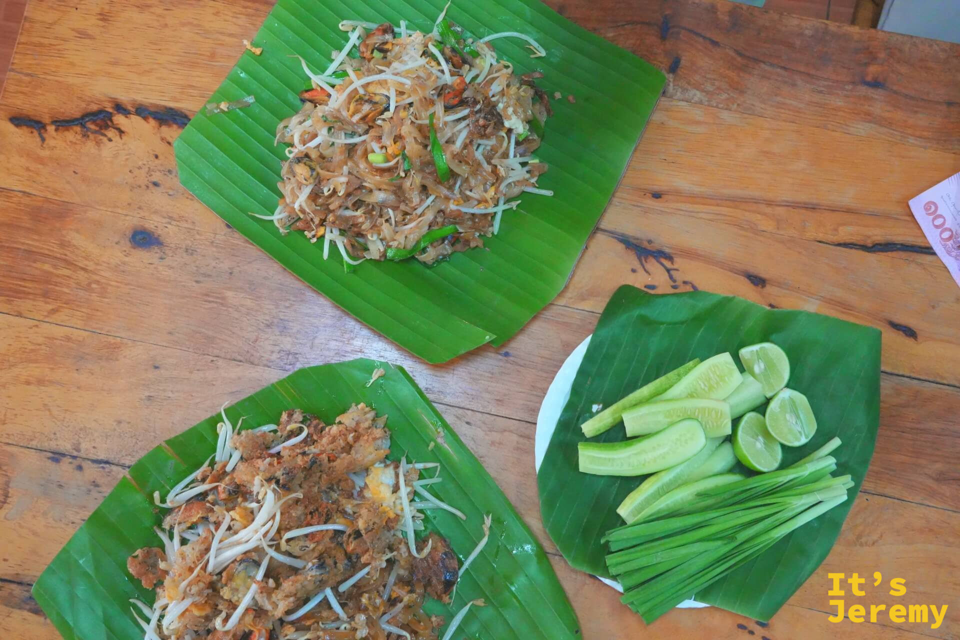 桌面鳥瞰：上方一份 Pad Thai、下方一份 Fried Mussel，右邊是配菜盤（小黃瓜、韭菜、萊姆），三樣全放在芭蕉葉上