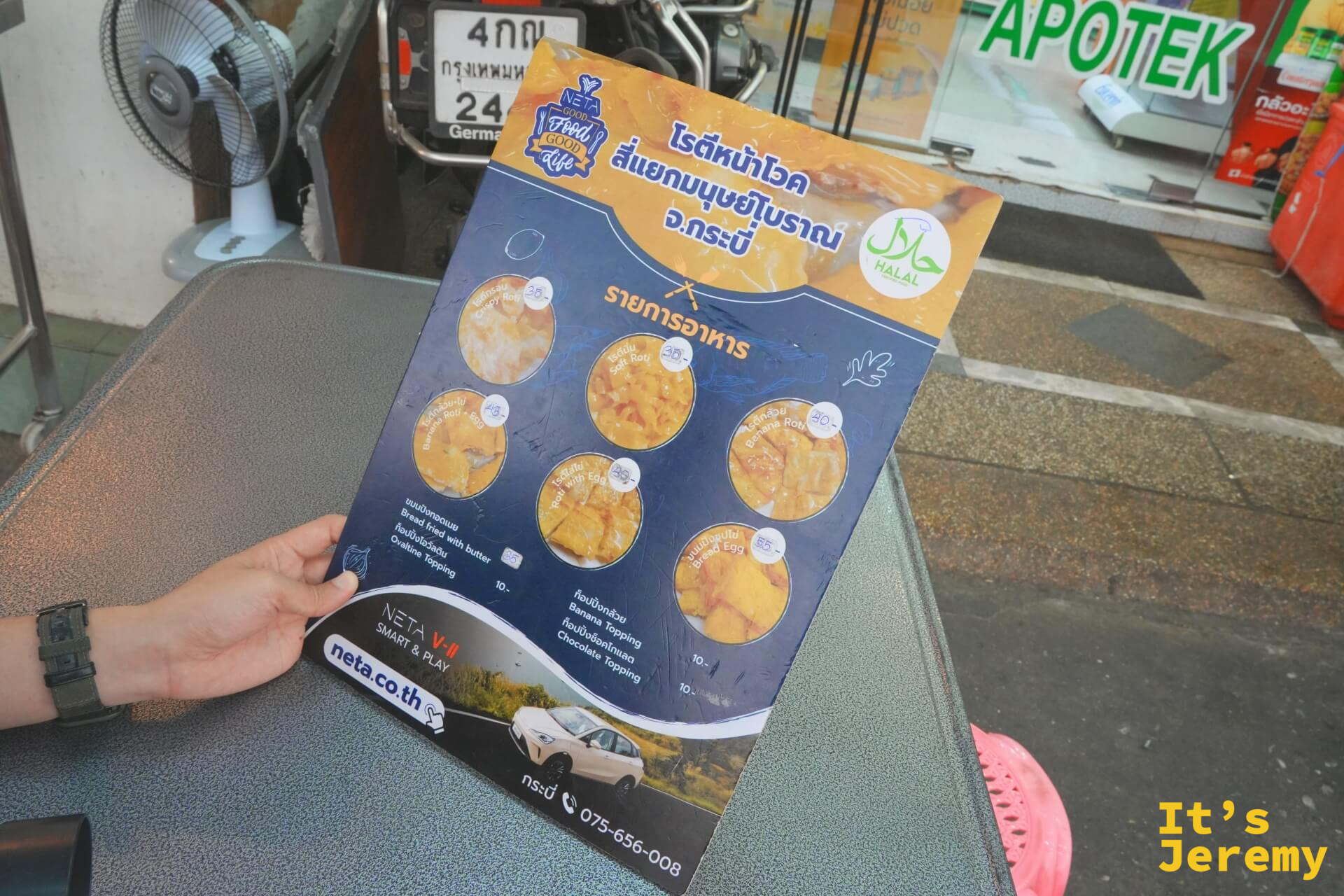 手持菜單，清楚列出各款 Roti 選項、Halal 認證標誌，以及 NETA 汽車廣告背面——典型喀比路邊攤的視覺混搭。
