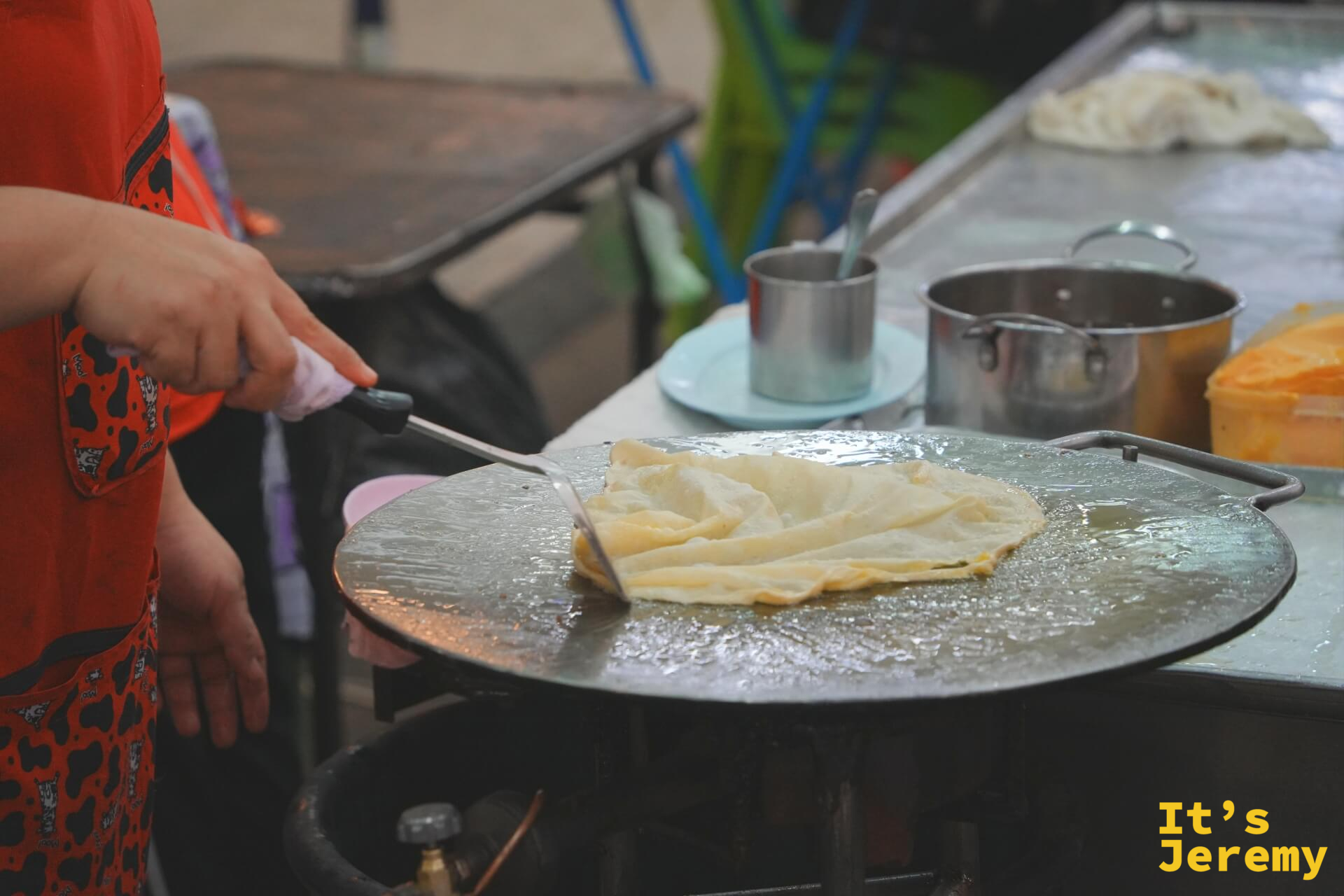 師傅在大圓鐵板上煎著一張薄透的千層 Roti 麵皮，麵皮邊緣呈現焦黃色。