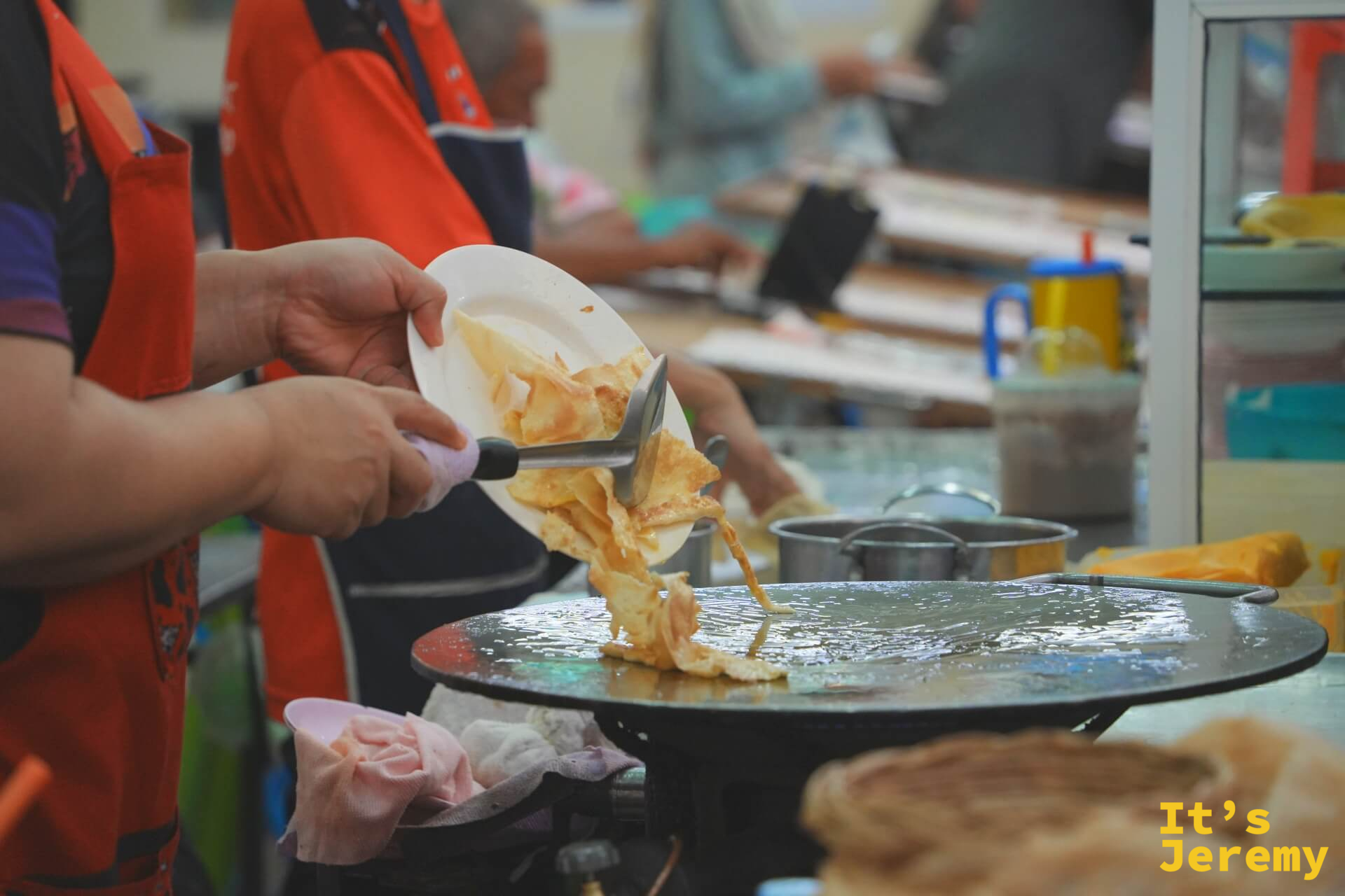 師傅將煎好的千層 Roti 用鏟子鏟起放入盤中，飛濺的油光在盤邊。