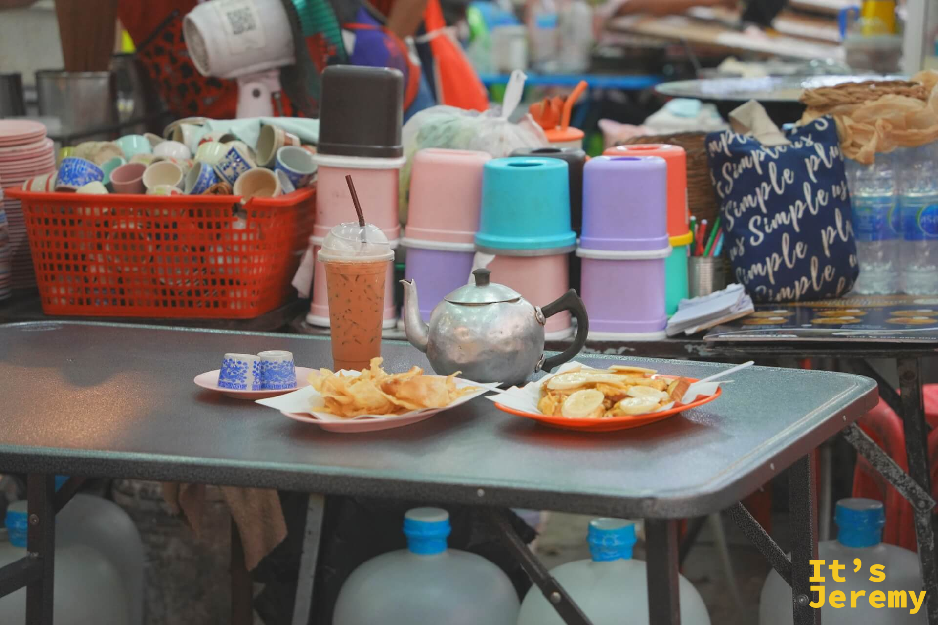 攤前桌面上擺著兩盤 Roti 點心、一壺鐵製熱茶壺和一杯冰奶茶，背景是疊放整齊的各色塑料杯與茶具籃。